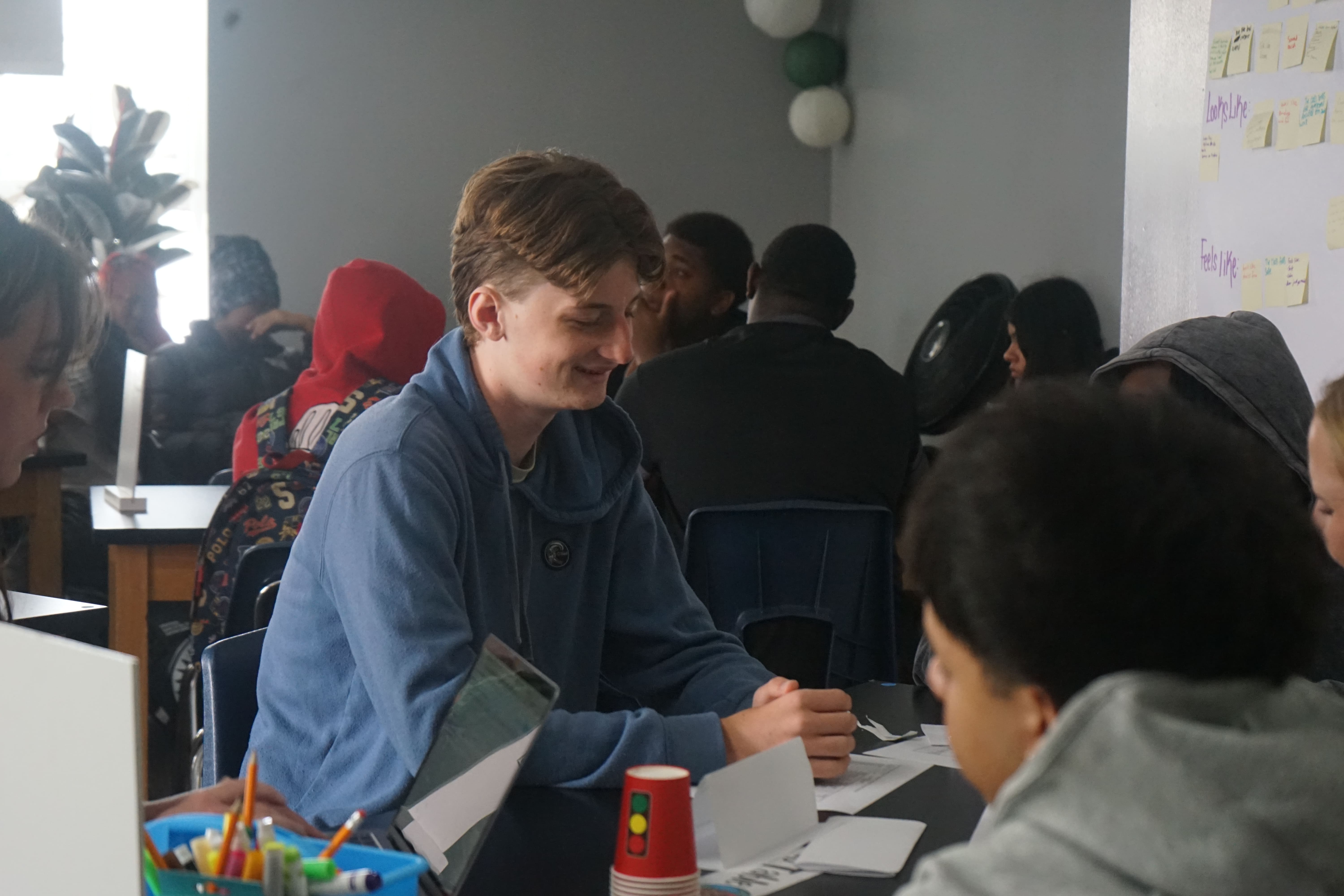 Two men working on an architectural model at a table in a bright room, one smiling at the camera while the other holds a model piece.