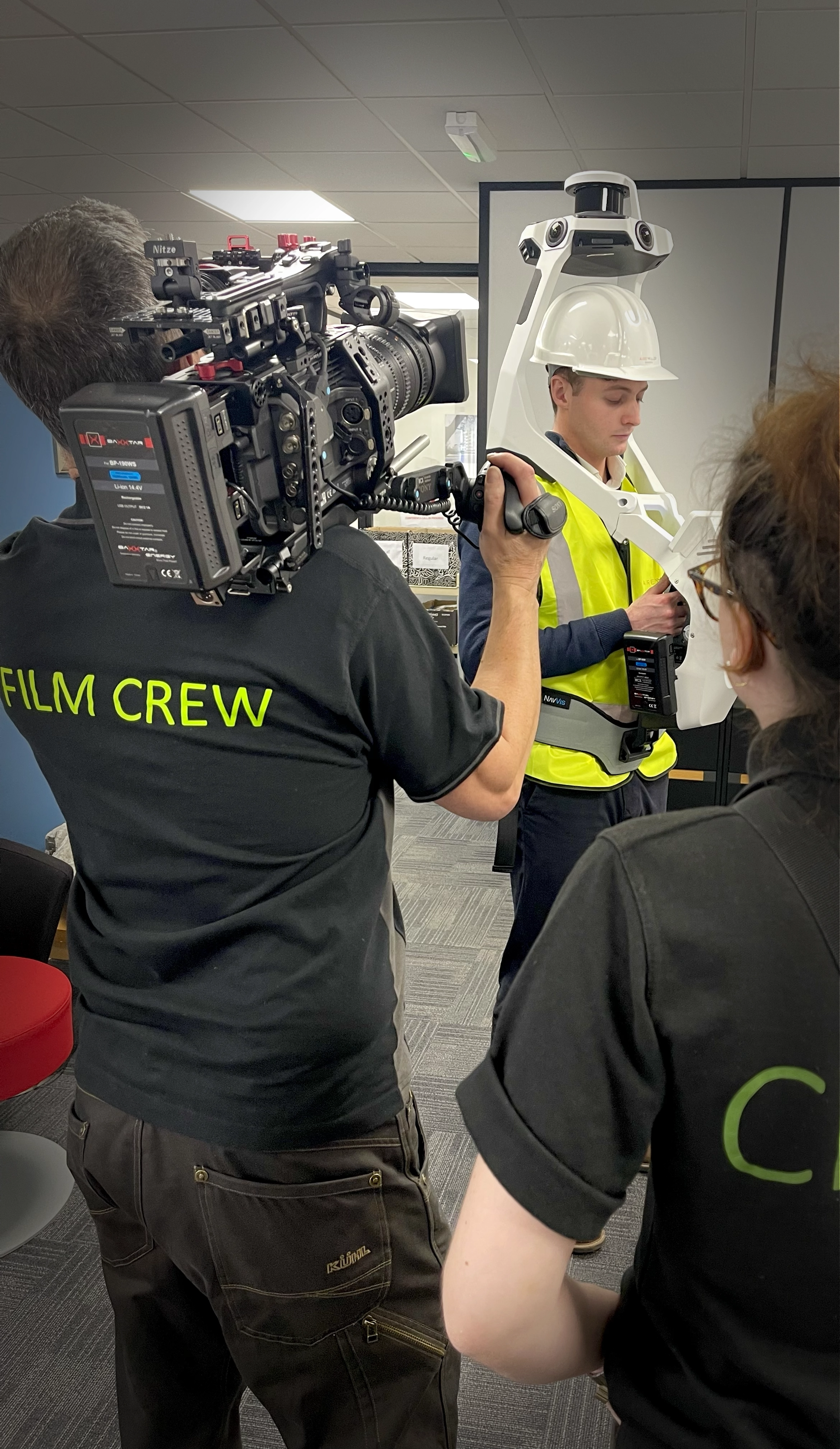 Woman wearing a green sweater holding a white mug and clipboard, standing in a creative workspace with a computer screen showing guitar video editing.