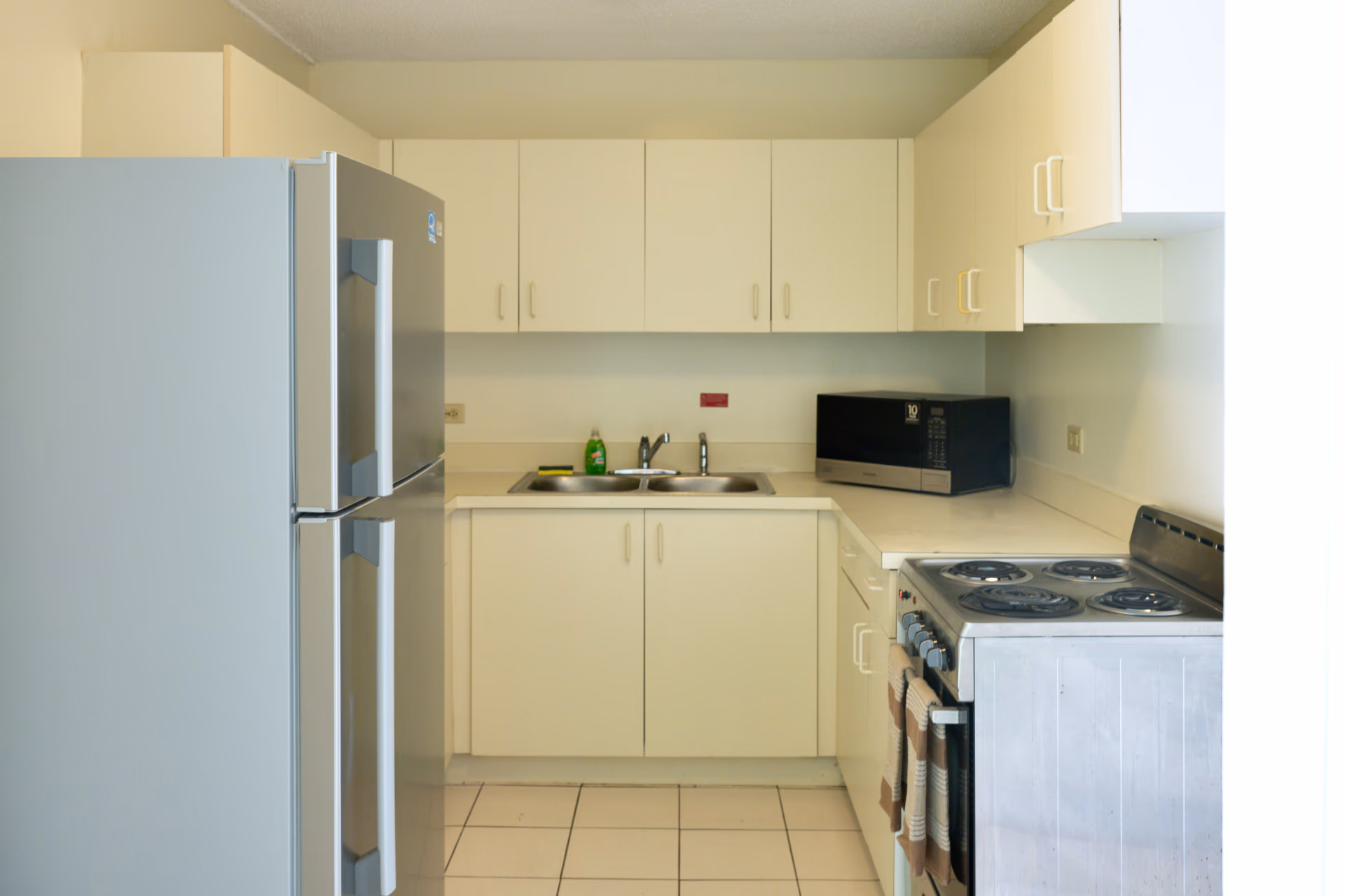 Compact kitchen with white cabinets, double sink, stainless steel refrigerator, microwave, and electric stove with towels.