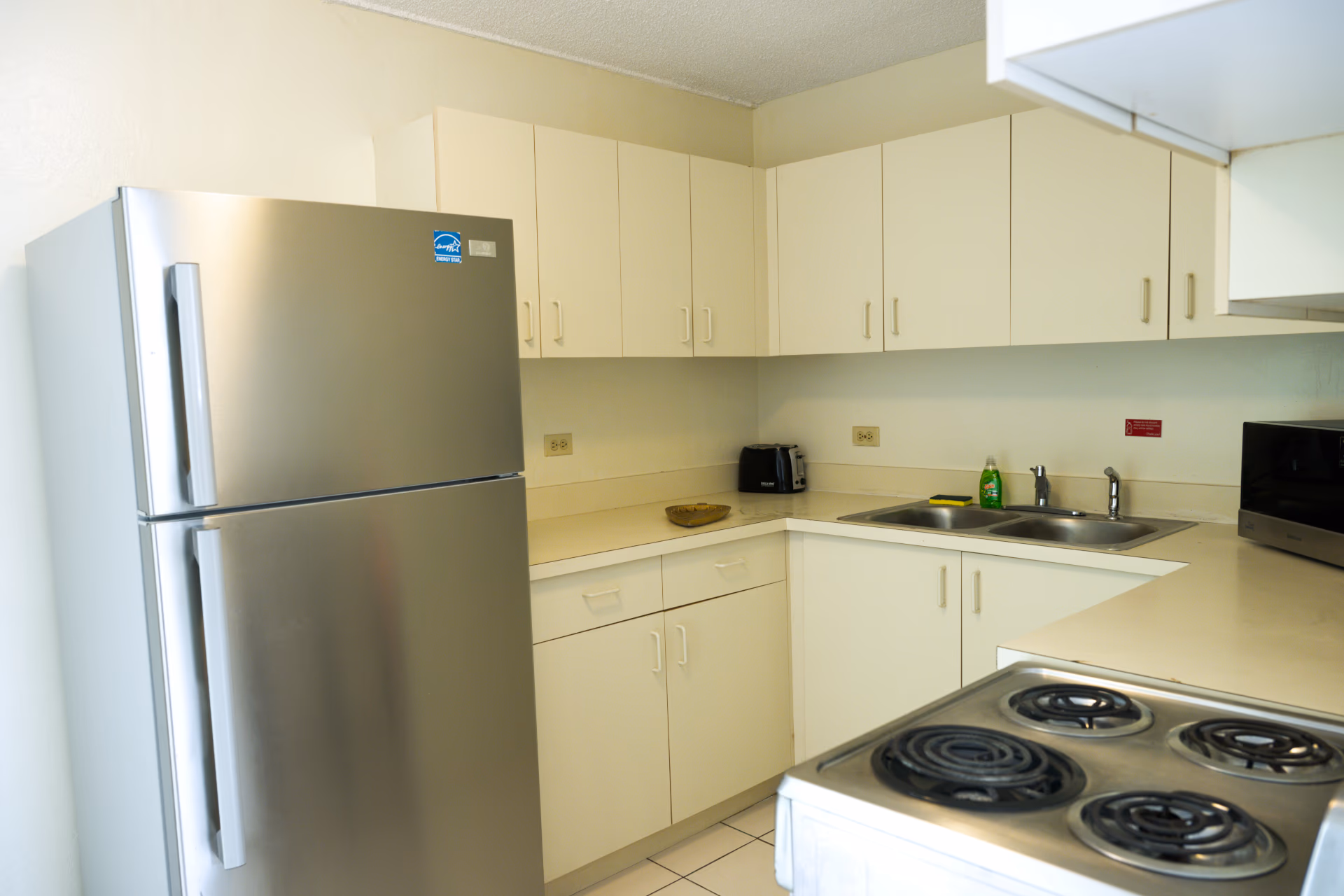 Clean kitchen corner with stainless steel refrigerator, white cabinets, double sink, electric stove, toaster, microwave, and dish soap.