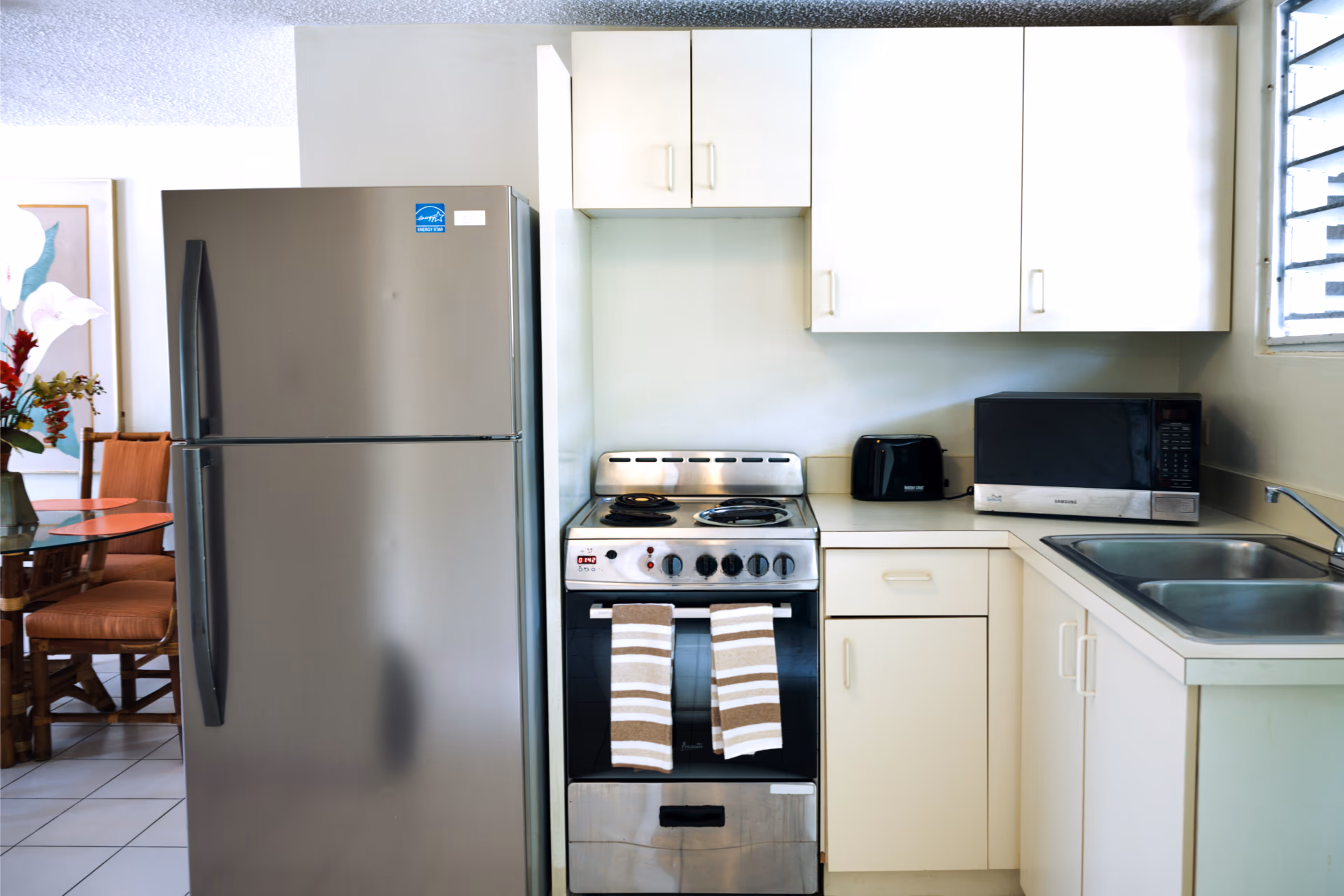 Compact kitchen with stainless steel refrigerator, stove with two striped towels, black toaster, microwave, and double sink.