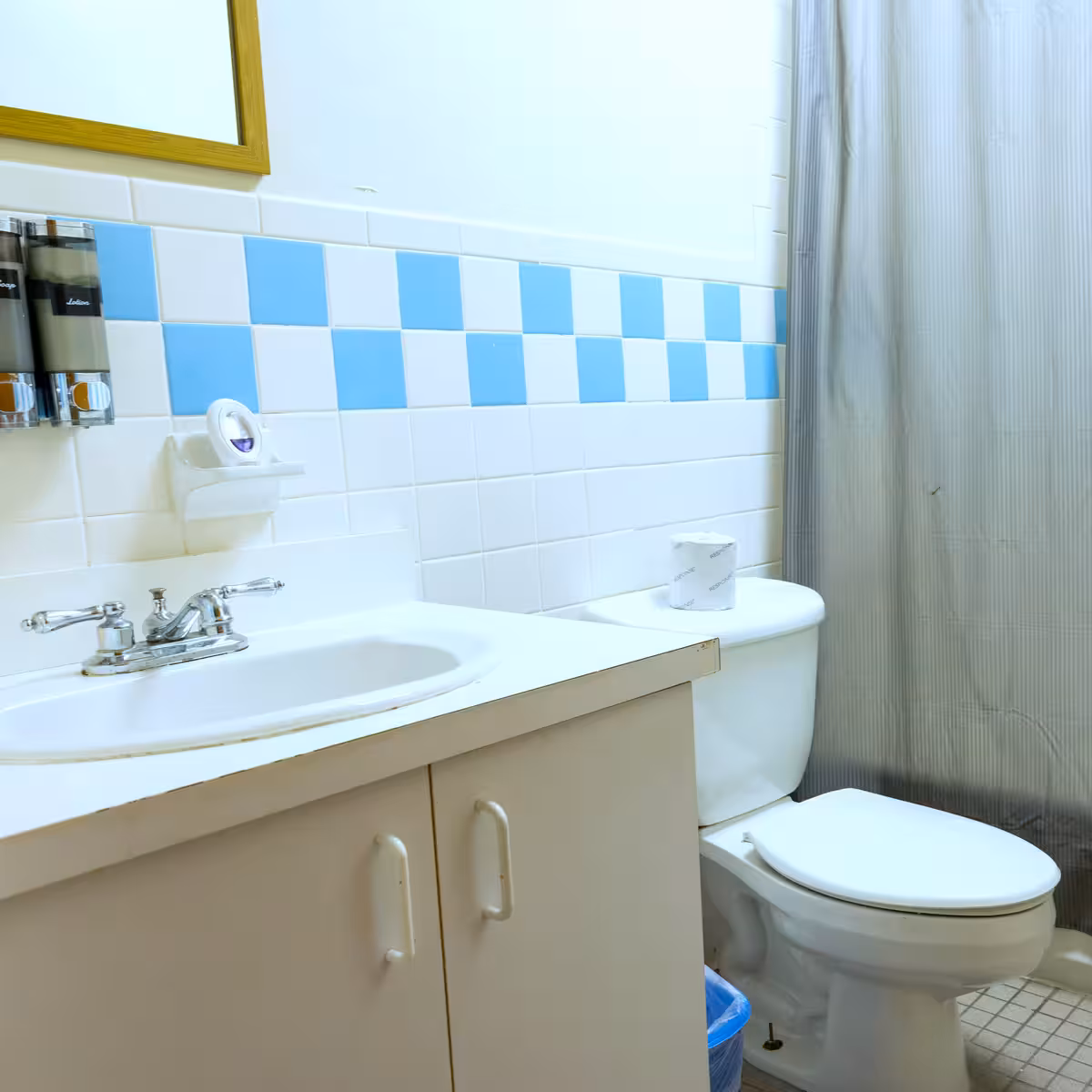 Bathroom with white sink countertop, blue and white checkered tile backsplash, toilet with a roll of toilet paper on top, and gray shower curtain.