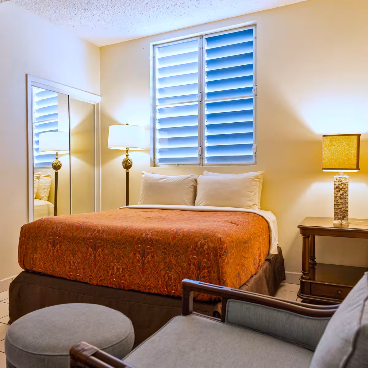 Bedroom with a bed featuring an orange patterned comforter, two white pillows, a tall lamp, a small side table with a square lampshade, a window with blue louvers, and a gray armchair with matching ottoman.