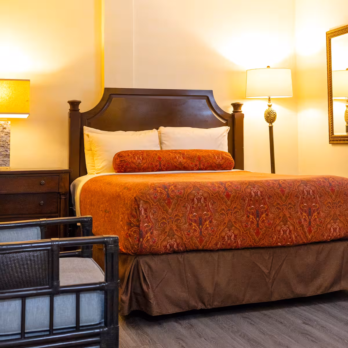 Hotel room with a bed featuring a dark wood headboard, two white pillows, an orange decorative bolster, and a patterned orange bedspread, flanked by a nightstand with a lamp on the left and a floor lamp with a mirror on the right.