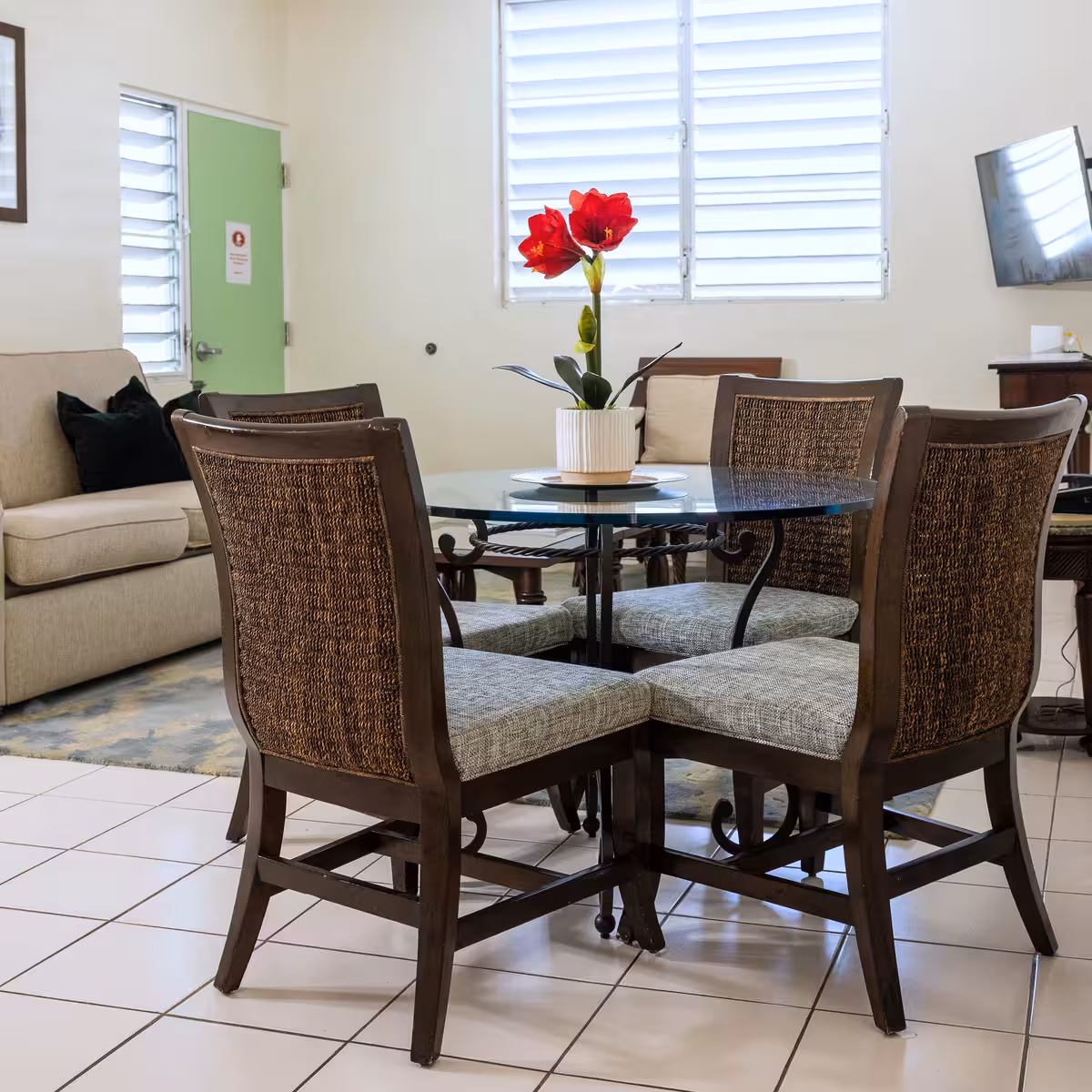 Dining area with a round glass table surrounded by four wooden chairs with woven backs and cushioned seats, featuring a white pot with red flowers on the table.