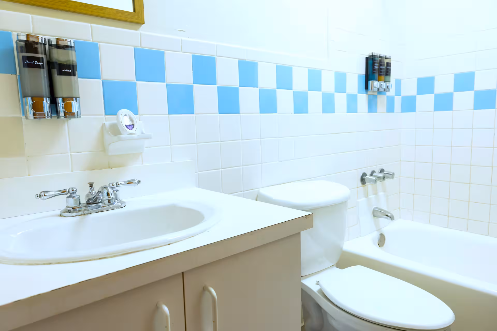 Bathroom with white sink and countertop, toilet, bathtub, and walls tiled in white and blue checkered pattern, with mounted soap and lotion dispensers.