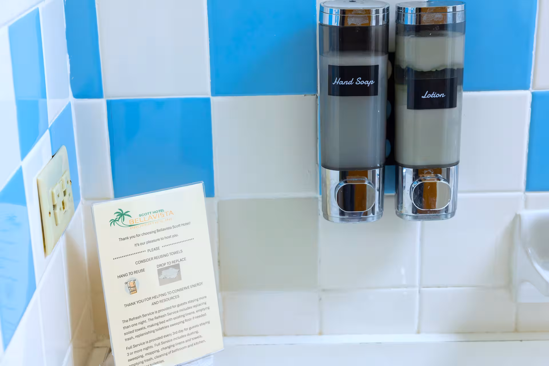 Bathroom wall with blue and white tiles, dual dispensers labeled Hand Soap and Lotion, and a notice about towel reuse from Bellavista Scott Hotel.