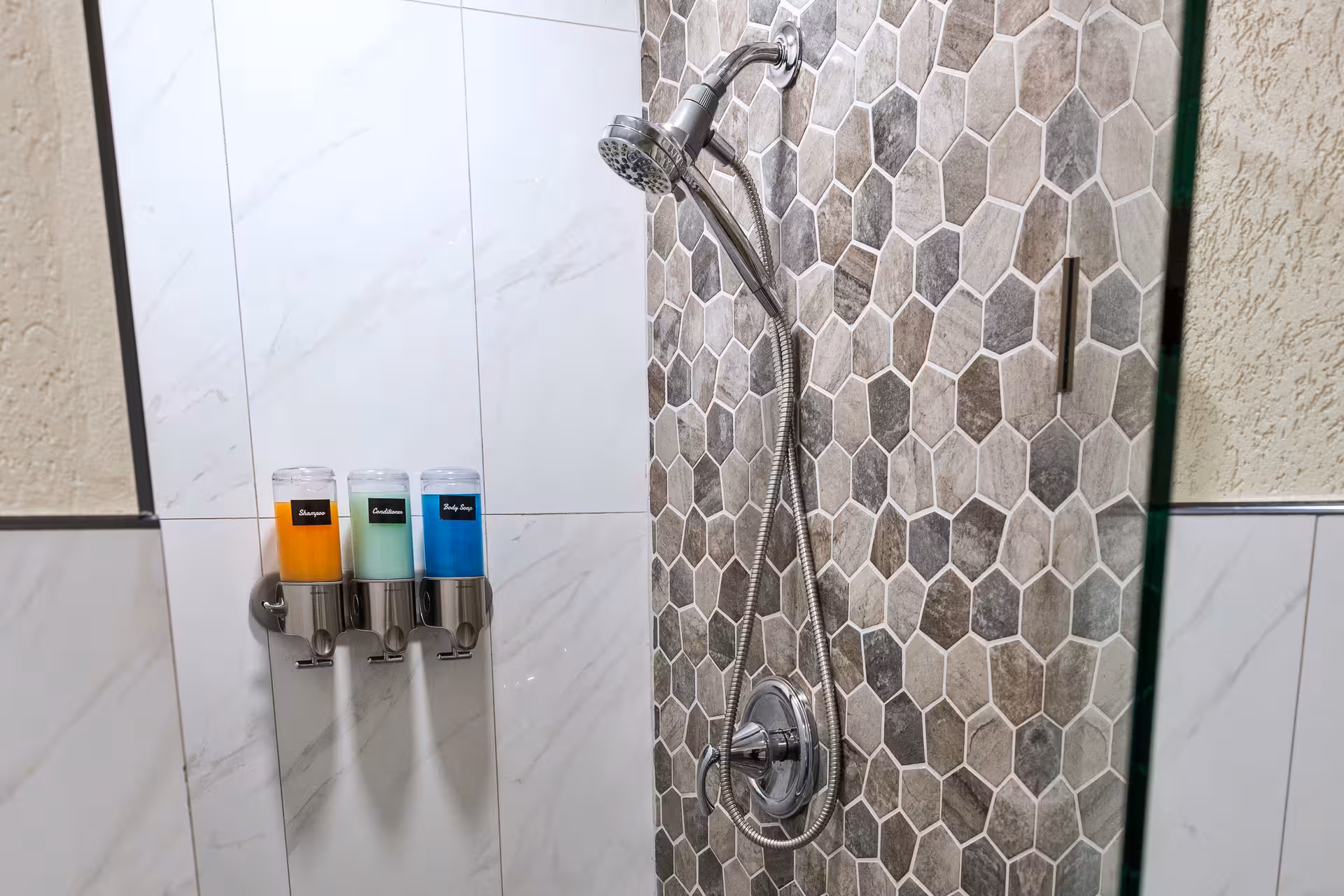 Shower area with hexagonal grey tile wall, chrome showerhead with flexible hose, and wall-mounted dispensers for shampoo, conditioner, and body soap.