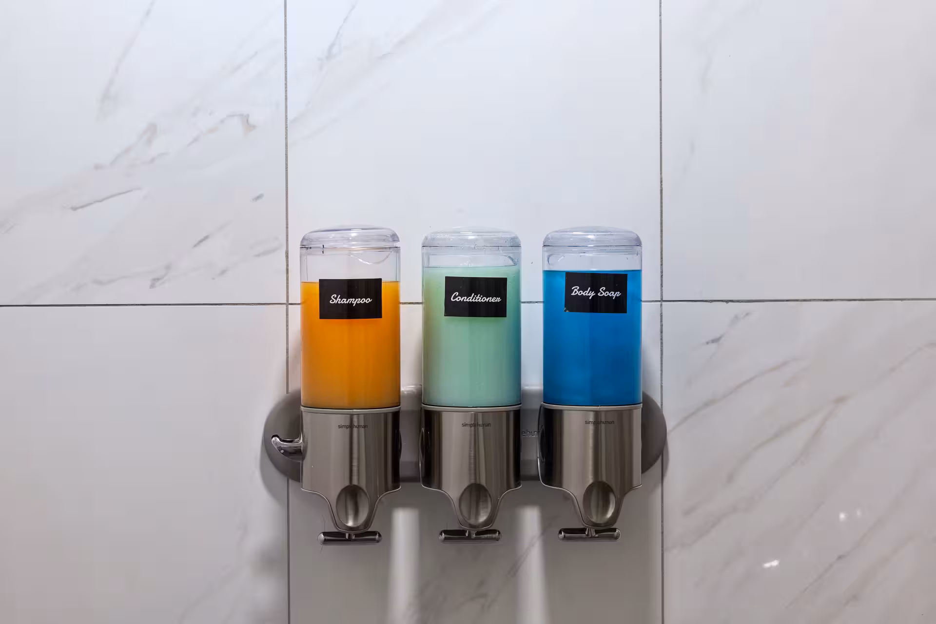 Wall-mounted dispensers with orange shampoo, green conditioner, and blue body soap against white tiled wall.