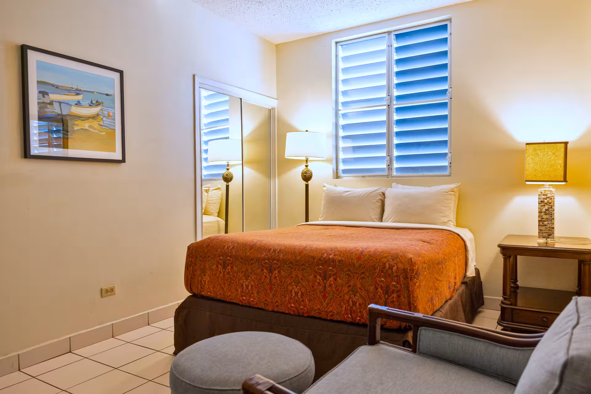 Cozy bedroom with a bed featuring orange patterned bedding, two pillows, a mirrored closet, window with shutters, and two lamps on either side.