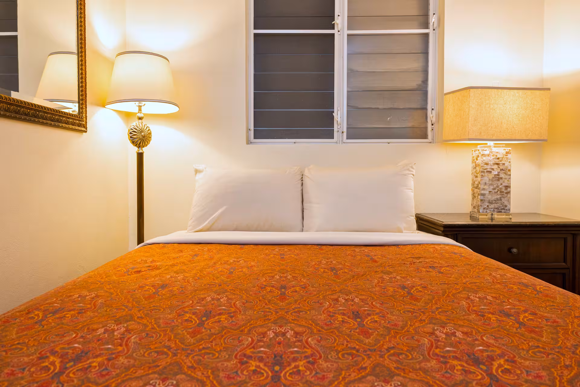 Bed with orange patterned blanket and white pillows, flanked by two lit lamps against a cream wall with a window and mirror.