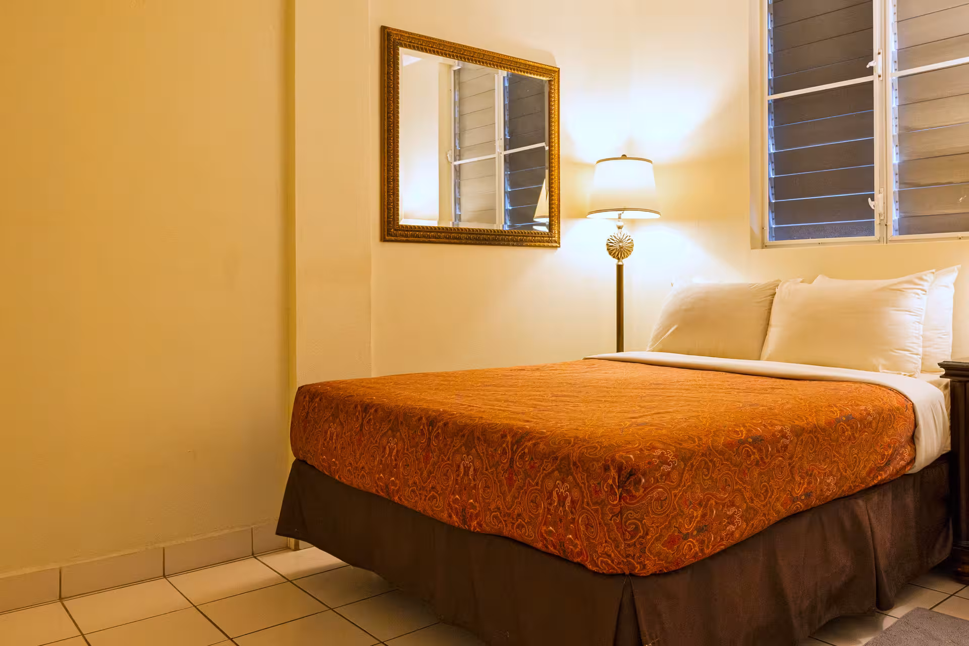 Bedroom with a bed covered in an orange patterned bedspread, white pillows, a floor lamp, and a window with horizontal blinds.