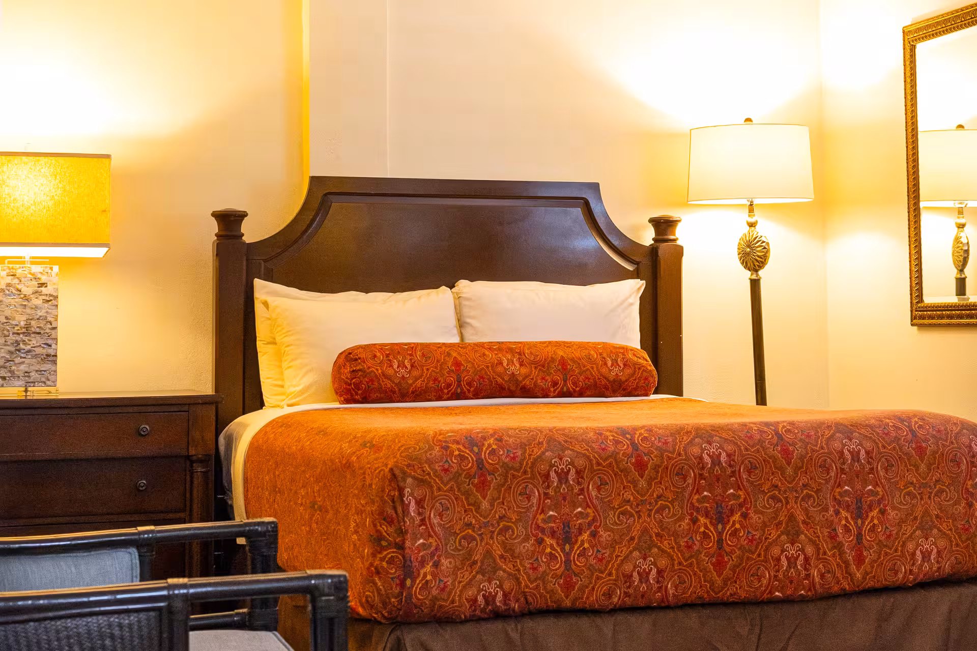 Hotel room with a large bed with a dark wooden headboard, orange patterned bedding, white pillows, and warm lighting from two lamps.
