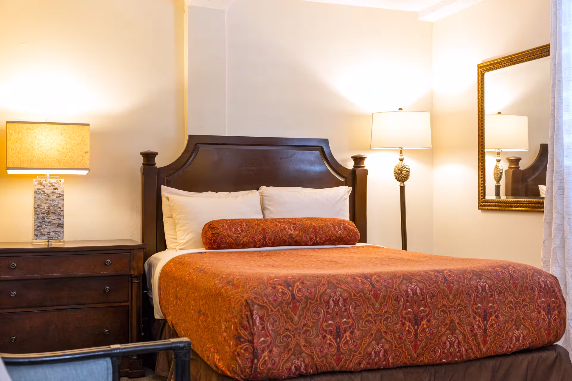 Cozy bedroom with dark wooden headboard, dresser with table lamp, patterned orange bedspread, bolstered pillow, floor lamp, and wall mirror.