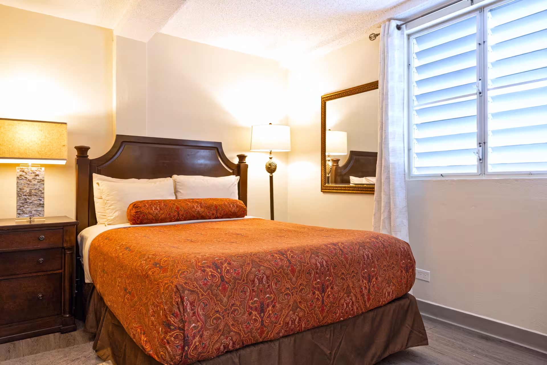 Bedroom with a wooden bed frame, orange patterned bedspread, two pillows, bedside table with lamp, standing lamp, and window with white curtains.