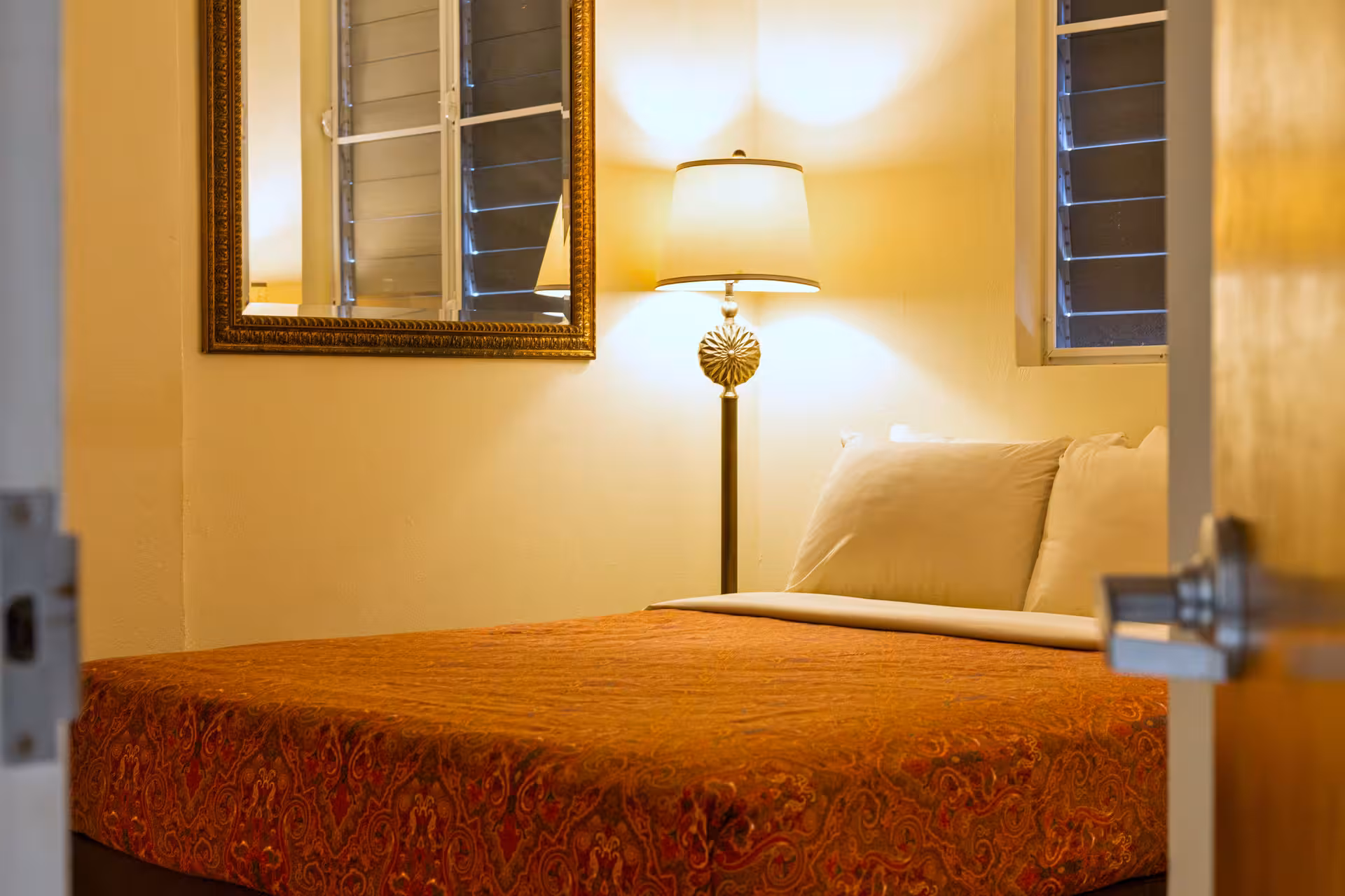 Cozy bedroom with a patterned orange bedspread, white pillows, a lamp on a bedside table, and a framed mirror on the wall.