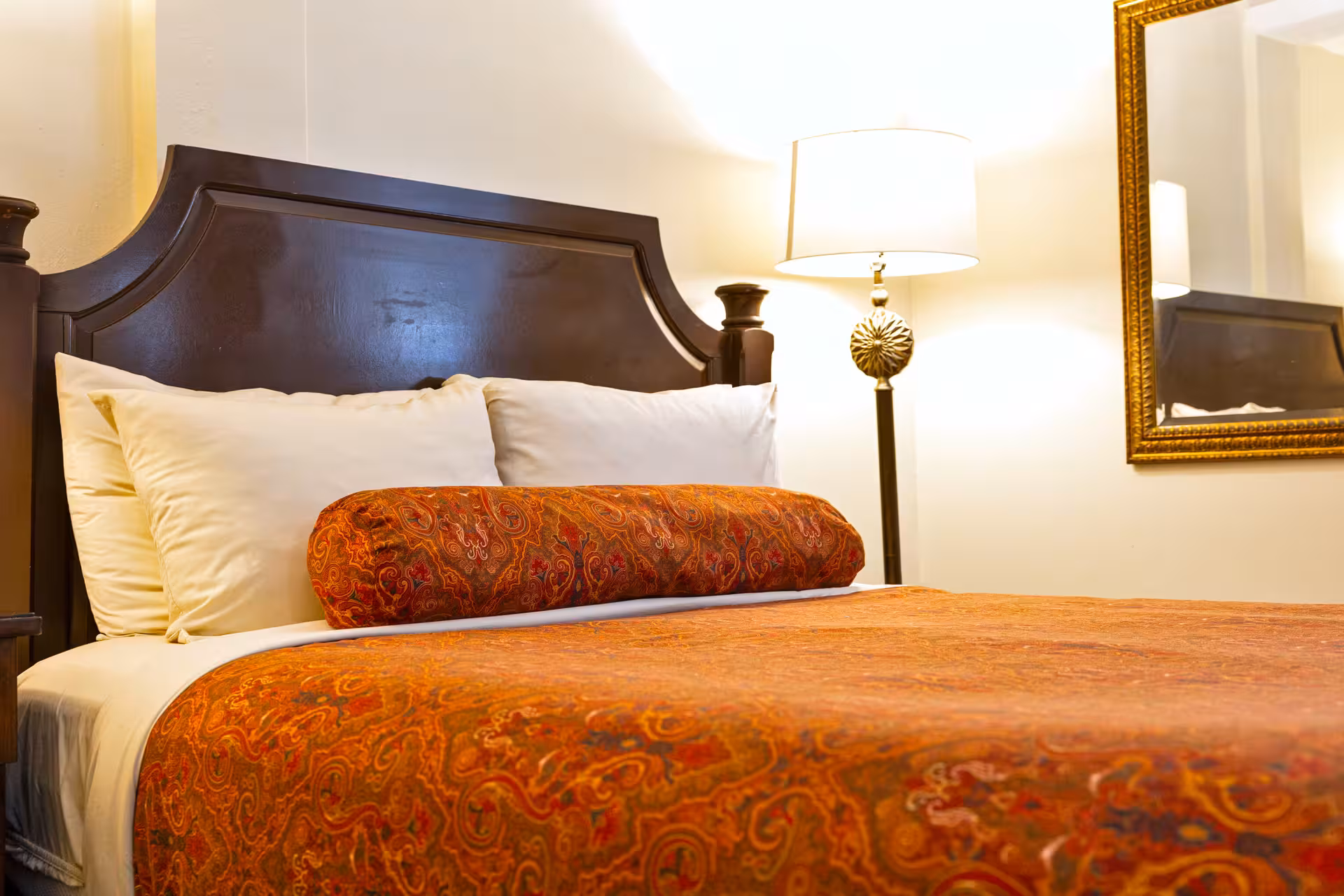 Bed with dark wooden headboard, white pillows, and orange paisley-patterned bedspread with matching bolster pillow, next to a lit floor lamp and a gold-framed mirror on the wall.