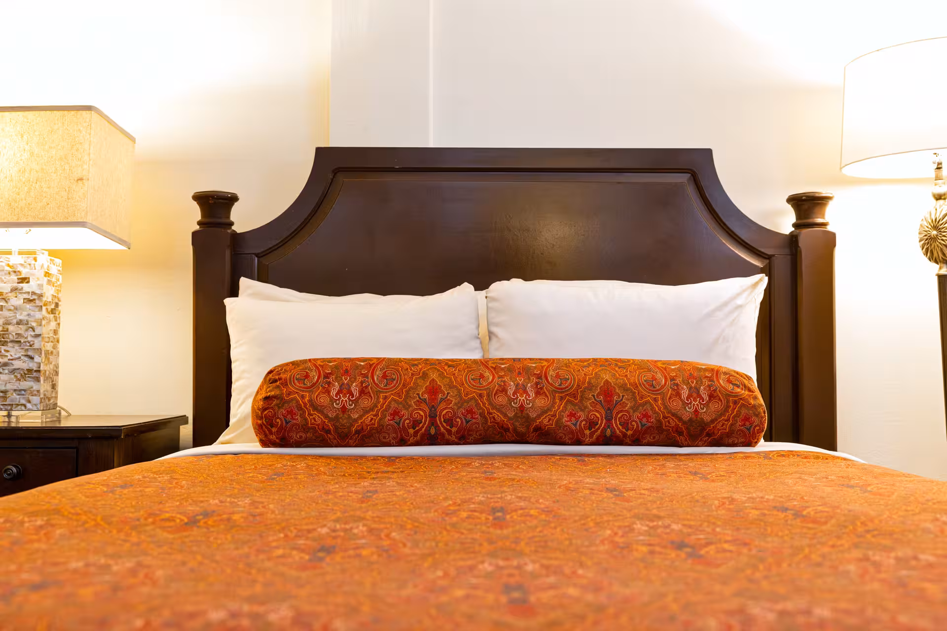 Bed with dark wooden headboard, white pillows, an orange patterned bolster pillow, and matching orange bedspread, flanked by two bedside lamps.