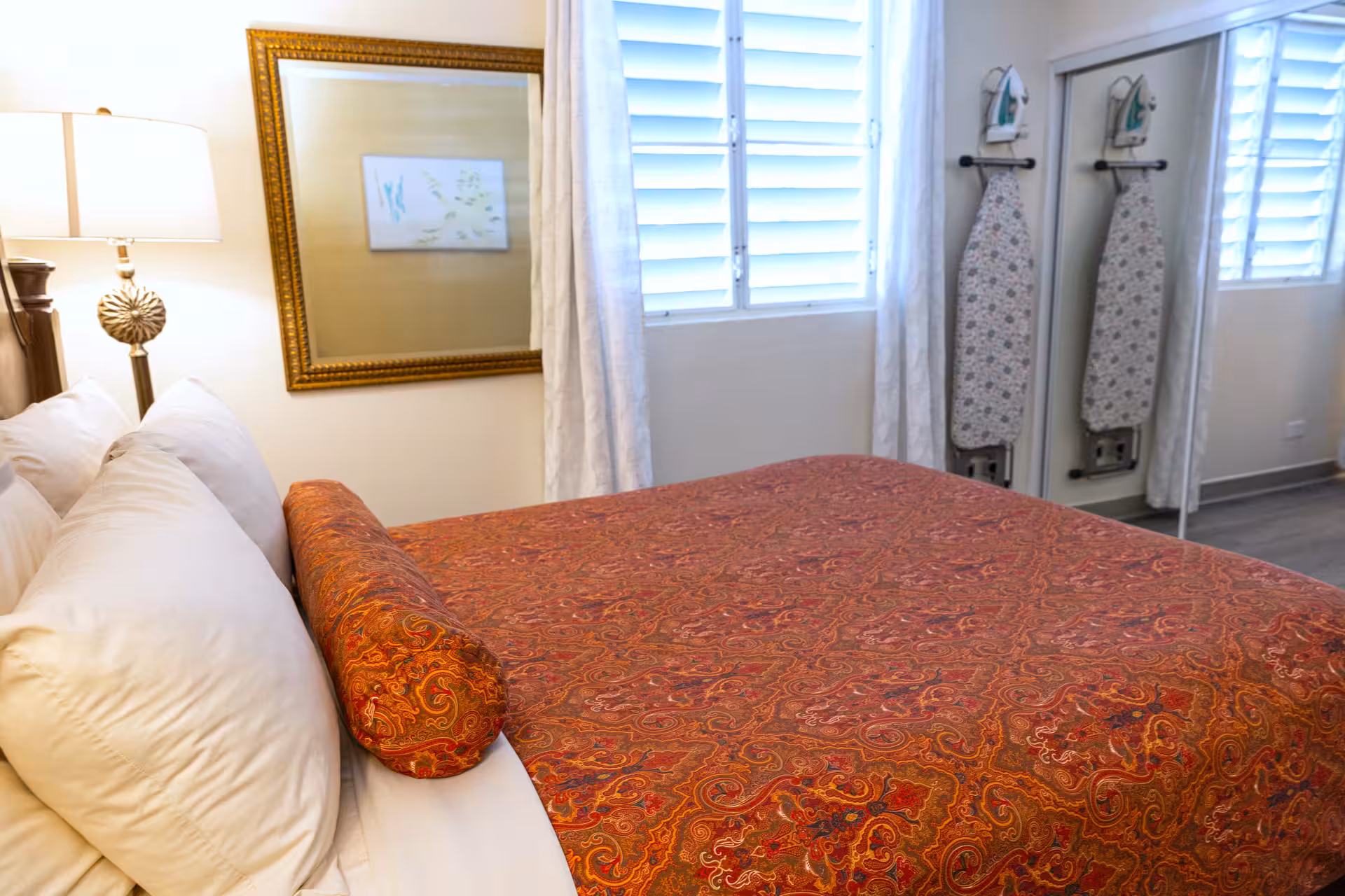 Bed with white pillows and a red patterned bolster pillow, next to a lit lamp and a gold-framed mirror.
