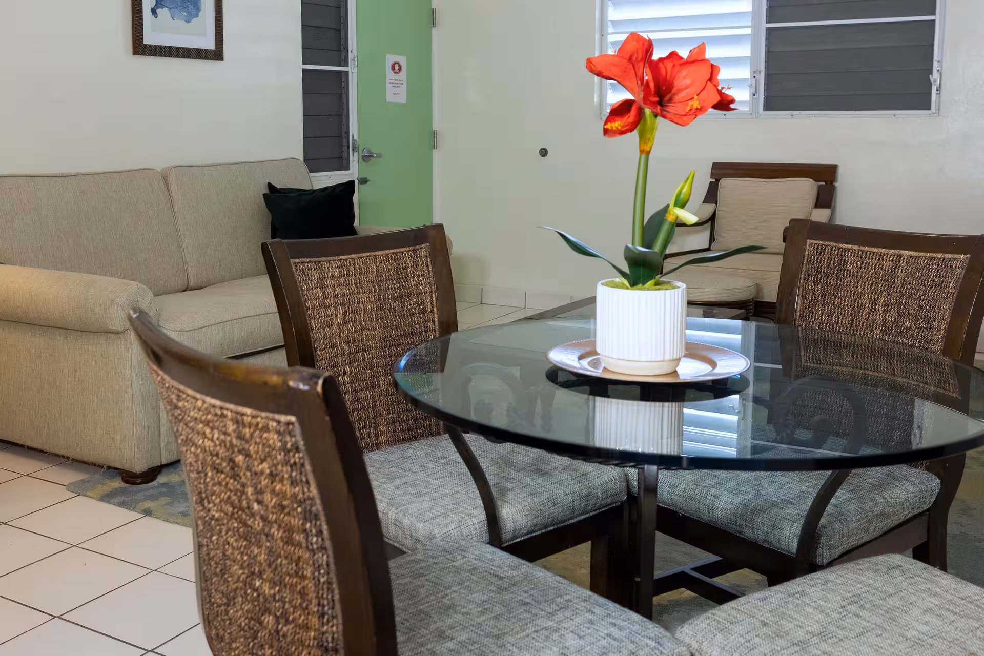 Modern dining area with a glass table, four cushioned chairs, and a red flower centerpiece in a white pot, with a beige couch and armchair in the background.
