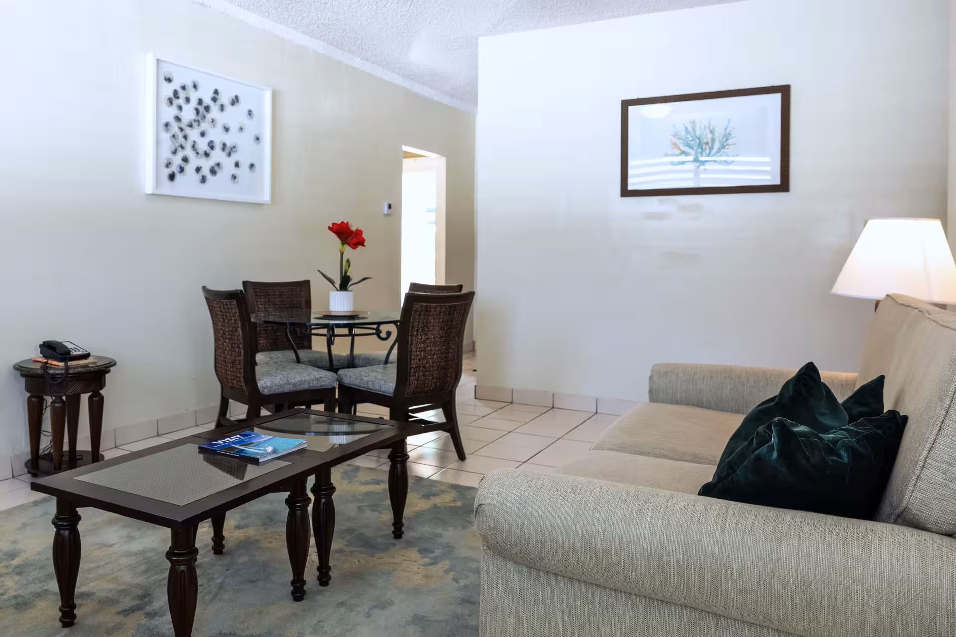 Living room with beige sofa and dark green pillows, glass coffee table, round dining table with four chairs and red flower centerpiece, and wall art.