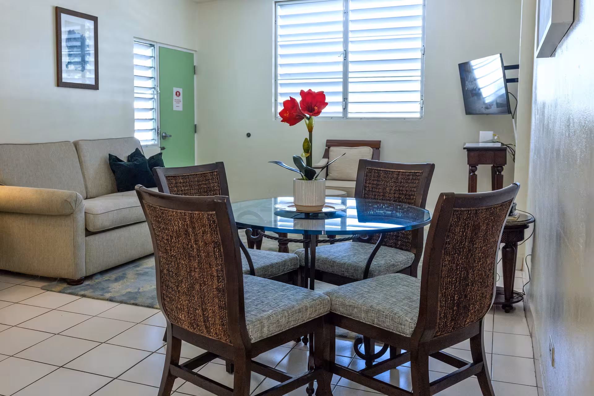 Modern living room with a beige sofa, glass dining table with four chairs, a red flower centerpiece, and a wall-mounted TV.