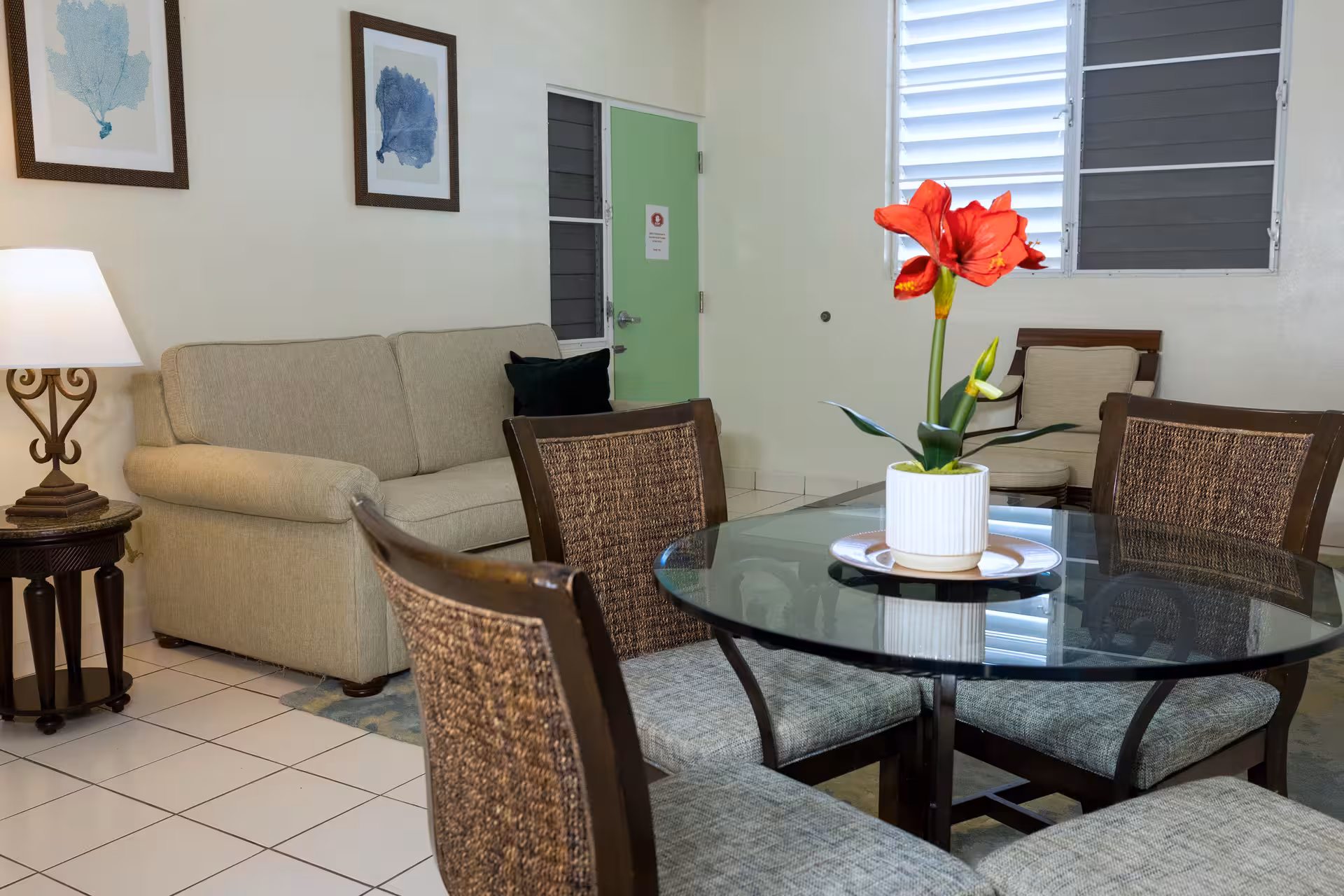 Living room with a glass dining table, four cushioned chairs, a beige sofa, armchair, side table with a lamp, and a red flower centerpiece on the table.
