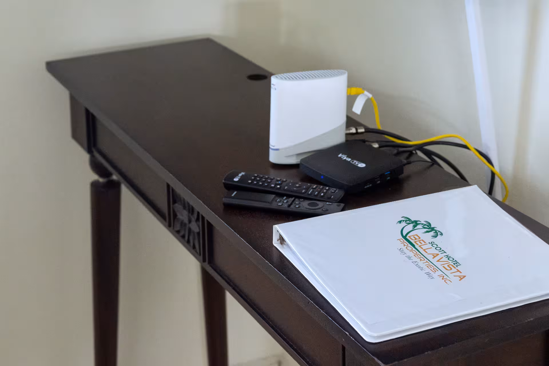Dark wooden desk with two remote controls, a white secure wifi router, a black Viva TV box, and a white binder labeled Scot Hotel Bellavista Properties Inc.