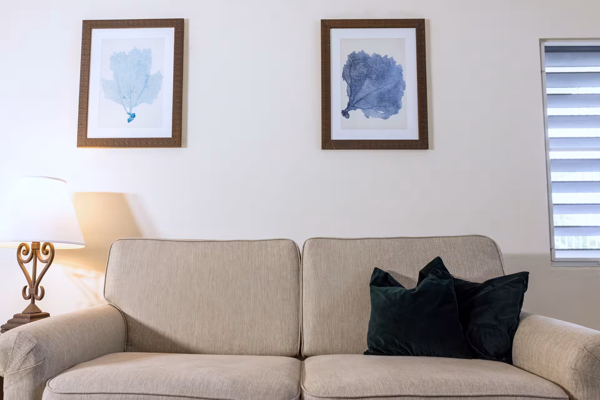 Beige sofa with two dark green pillows, a lamp on the left, and two framed blue coral prints on the wall.