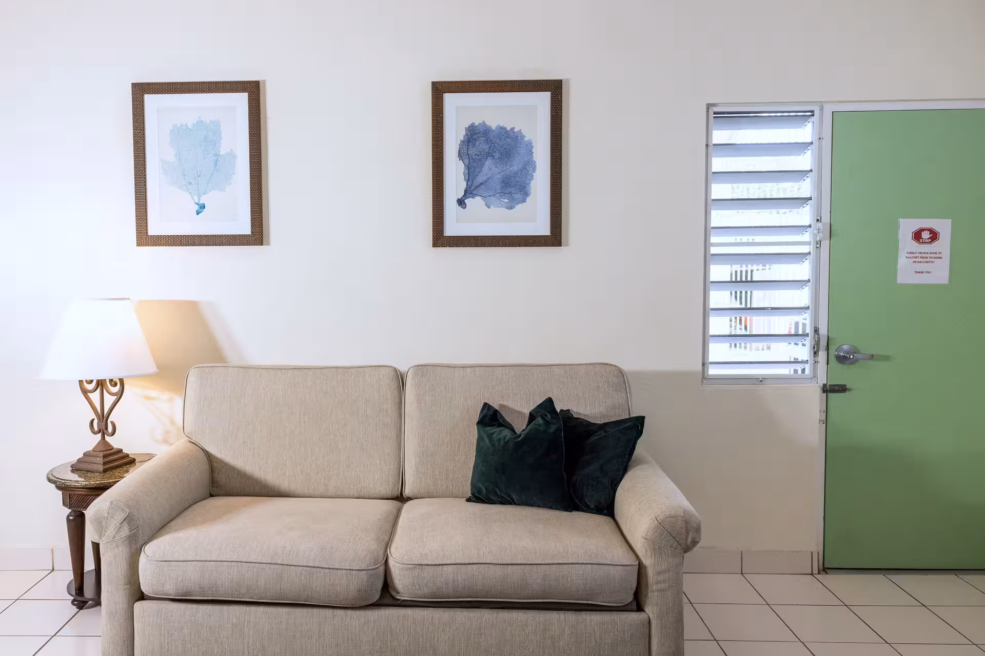 Beige loveseat with two dark green throw pillows centered under two framed blue coral prints, next to a side table with a lamp and a green door with a louvered window.