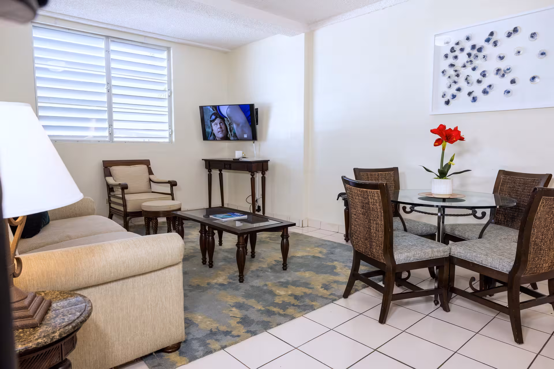 Living room with beige sofa, armchair, dark wood coffee table on patterned rug, wall-mounted TV, and round glass dining table with four chairs and red flower centerpiece.