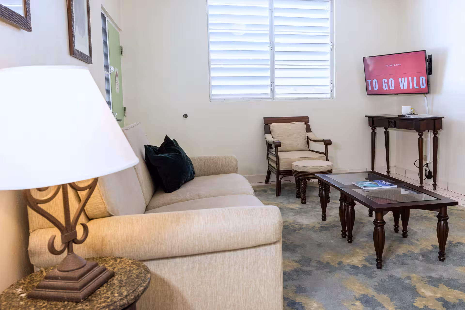 Living room with beige sofa, a wooden chair with cushion, a glass coffee table, a TV displaying 'TO GO WILD', and a table lamp.