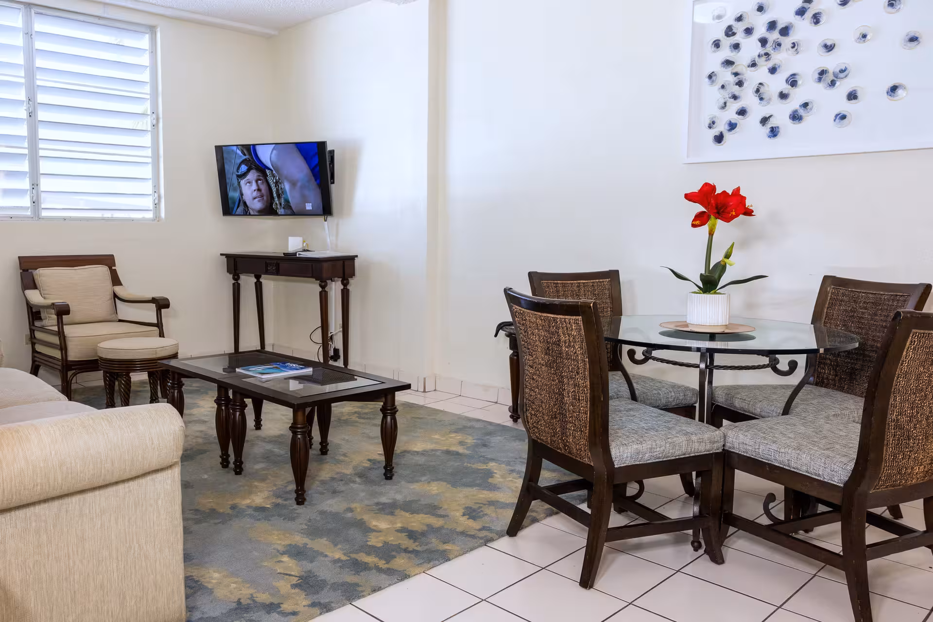 Living room with beige sofa, wooden armchair, glass coffee table, wall-mounted TV, and round glass dining table with four chairs and a red flower centerpiece.