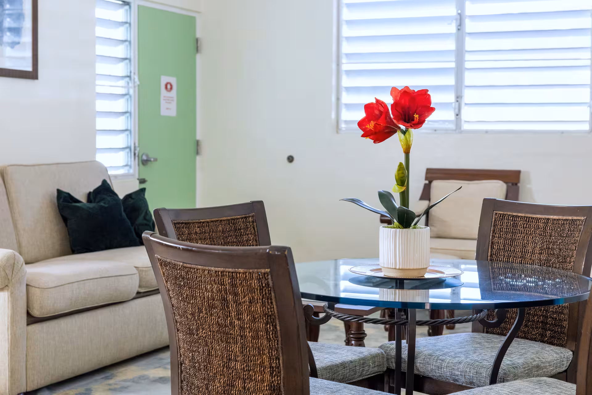 Dining area with a round glass table, four chairs, and a white pot holding a red flower centerpiece, next to a beige sofa with dark cushions.