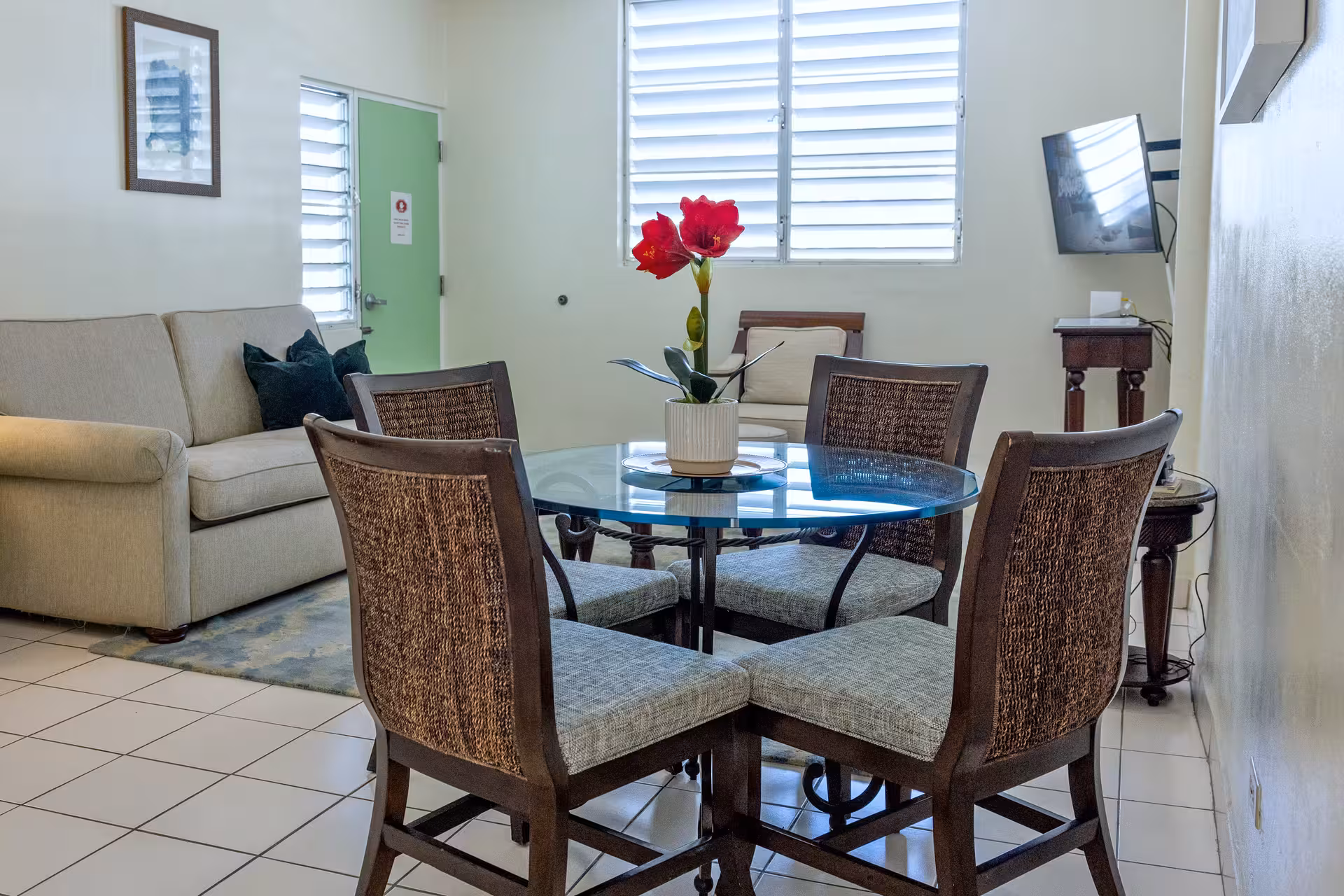 Dining area with a round glass table surrounded by four cushioned chairs with woven backs, a beige sofa with dark pillows, and a small wooden table with a mounted TV in a bright room.