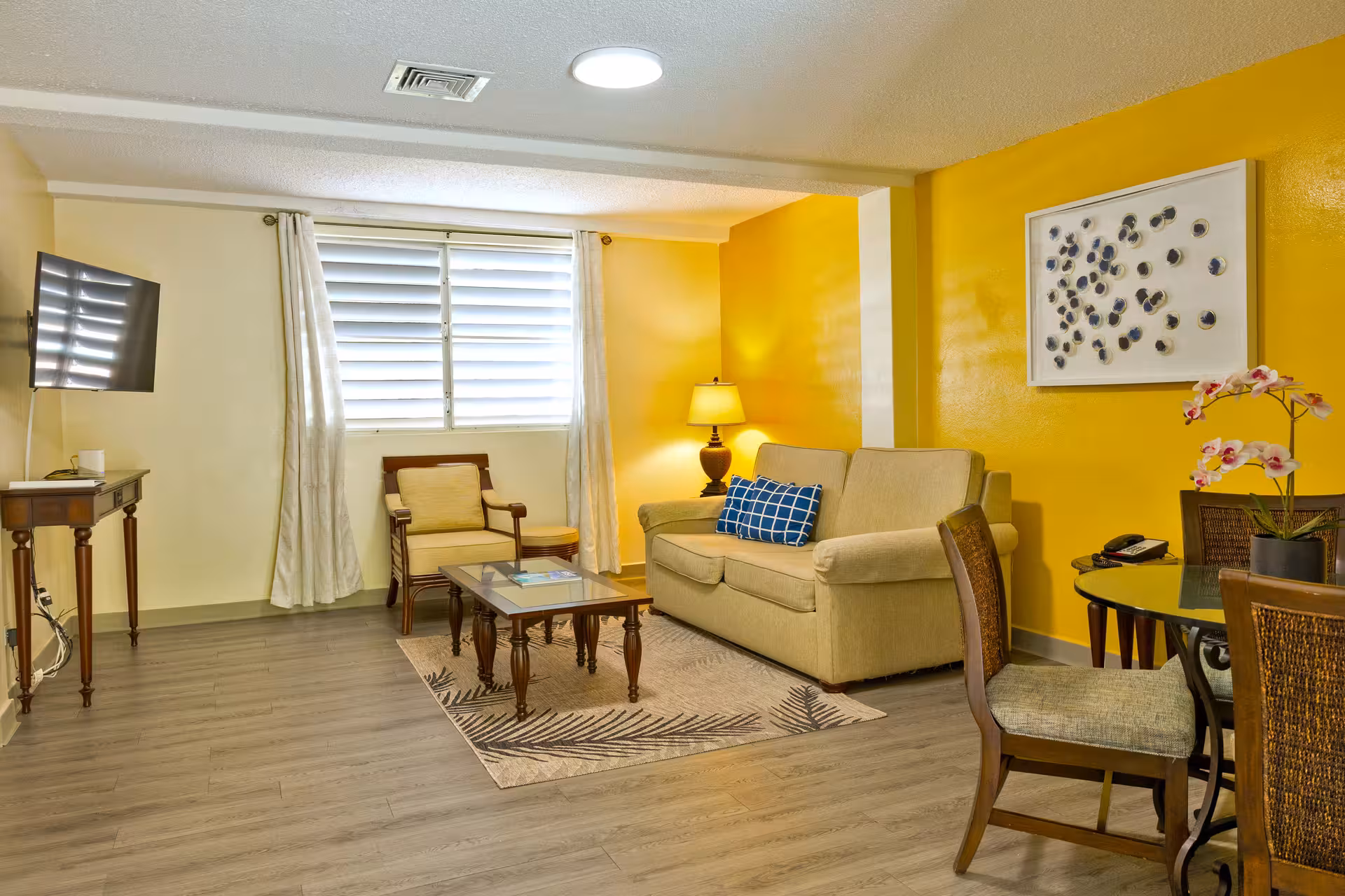 Living room with yellow accent wall, beige sofa with blue pillow, armchair, glass coffee table, wall-mounted TV, and dining table with chairs and orchid flower.