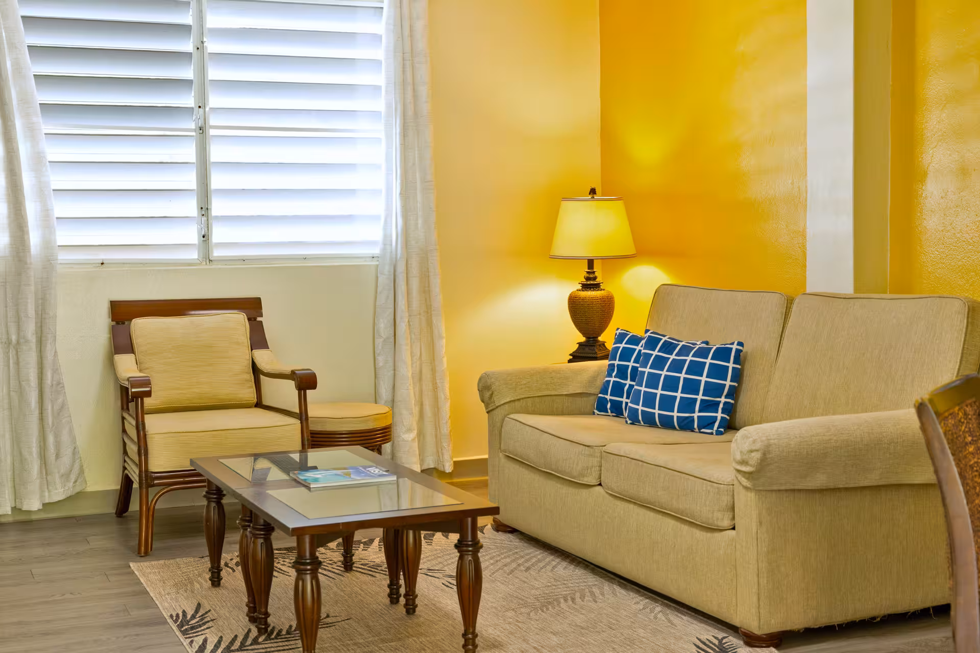 Cozy living room with a beige sofa, two blue patterned cushions, a matching armchair, a wooden coffee table, and a lamp against a yellow wall.