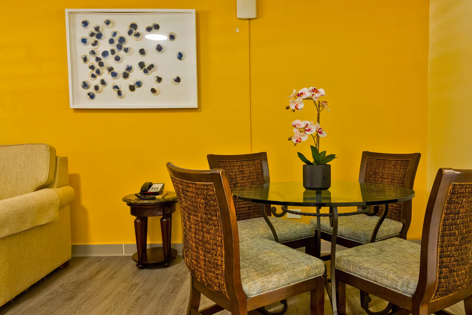 Dining area with a round glass table, four cushioned chairs with woven backs, a potted orchid centerpiece, and a yellow wall with abstract art.