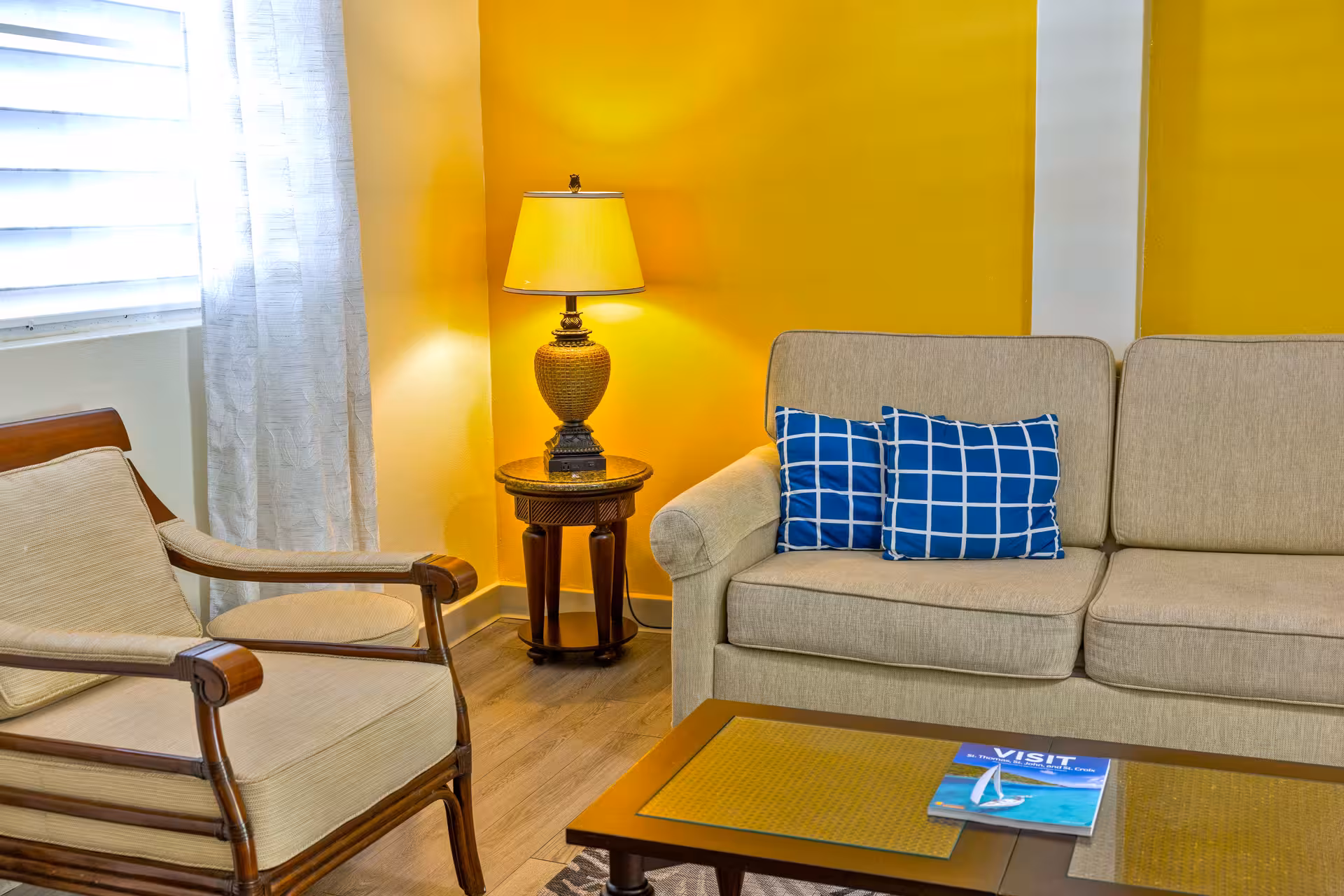 Living room corner with beige armchair and sofa with blue checkered pillows, wooden side table with lamp, and wooden coffee table with a magazine.