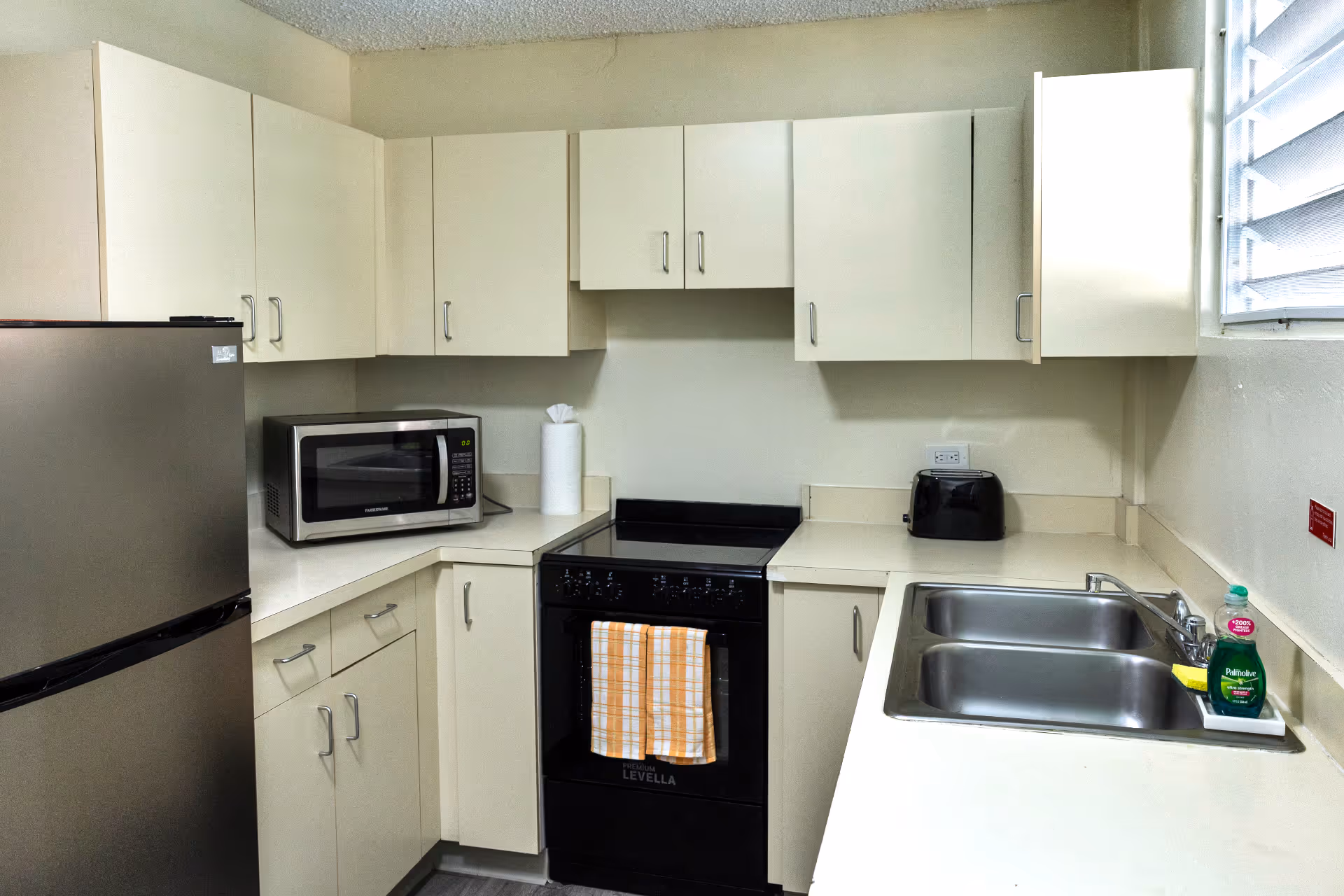 Compact kitchen with beige cabinets, black electric stove with a yellow checkered towel, stainless steel microwave, double sink, refrigerator, black toaster, and dish soap near window.
