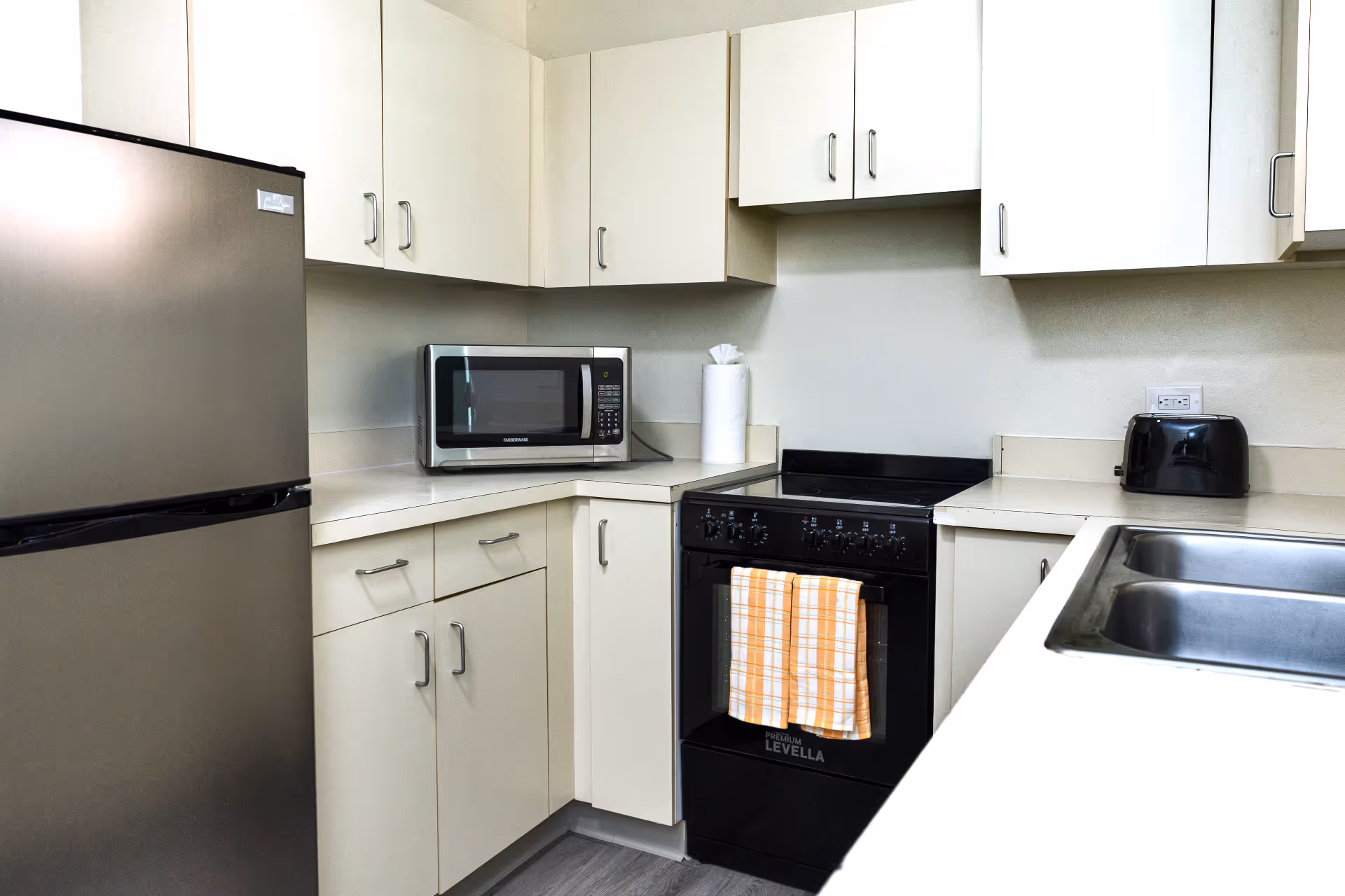 Modern kitchen corner with stainless steel refrigerator, microwave, black stove with orange and white checkered towels, black toaster, and double sink.