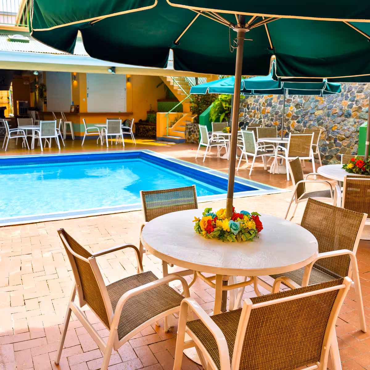 Outdoor pool area with round tables, chairs, green umbrellas, and a floral centerpiece on a table.