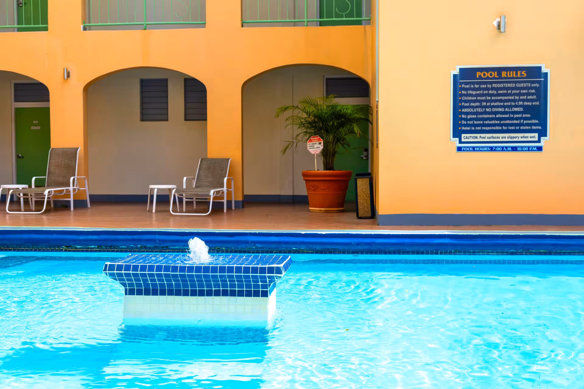 Outdoor swimming pool with a small blue tiled fountain, lounge chairs, and a wall displaying pool rules.
