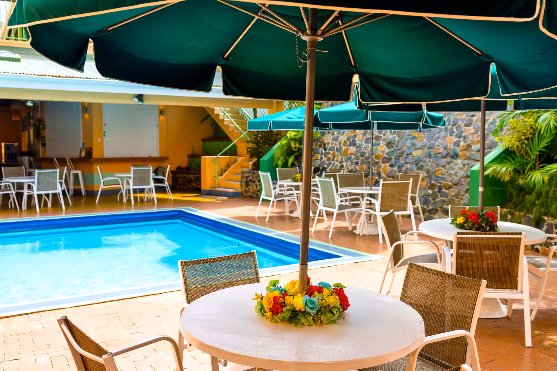 Outdoor pool area with several round tables, wicker chairs, large green umbrellas, and colorful floral centerpieces.