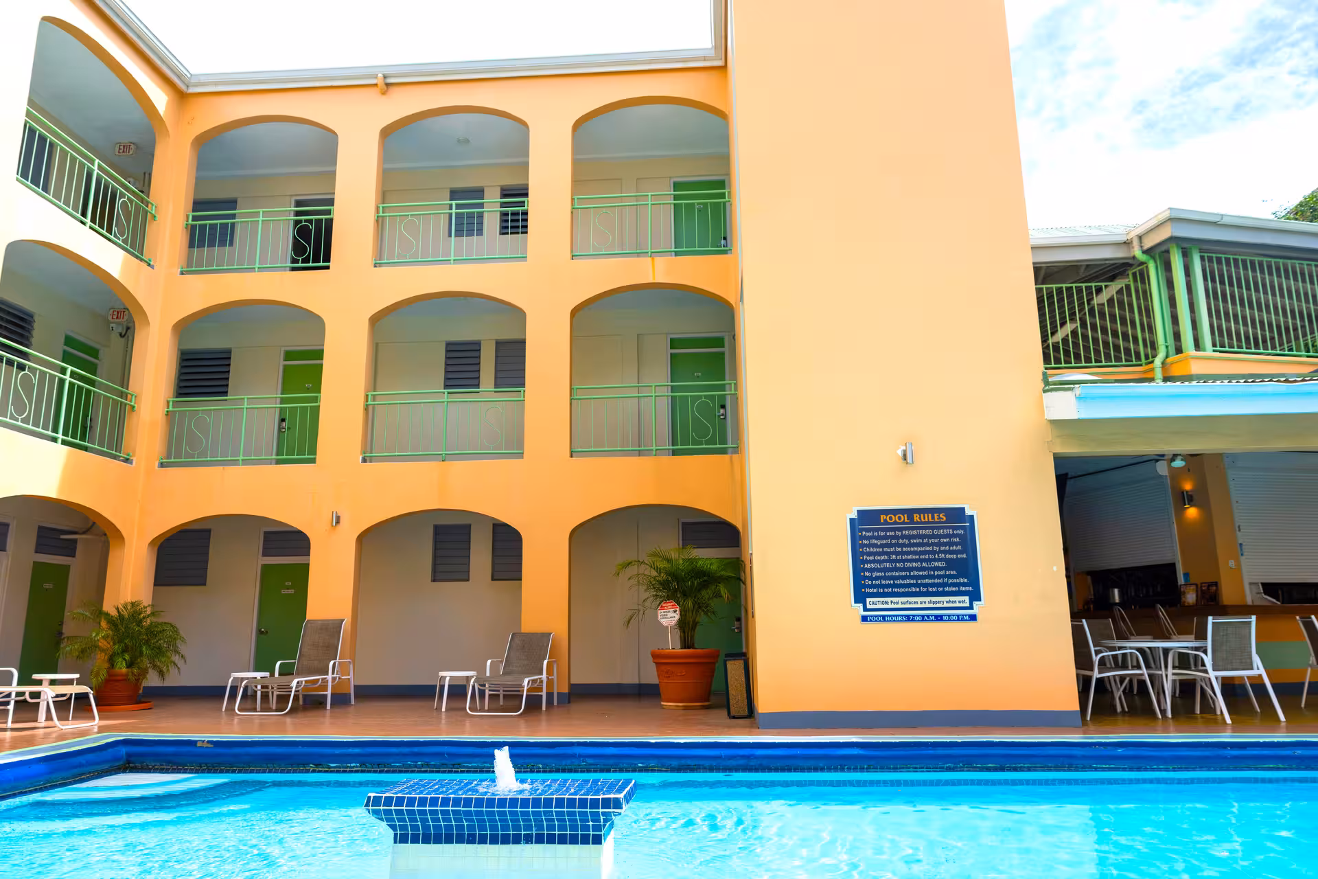 Outdoor hotel pool with lounge chairs, potted plants, and three-story building with green doors and railings in the background.