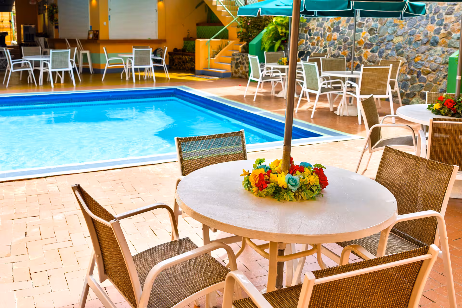 Outdoor seating area with white tables and brown chairs around a swimming pool, decorated with a colorful floral centerpiece under an umbrella.