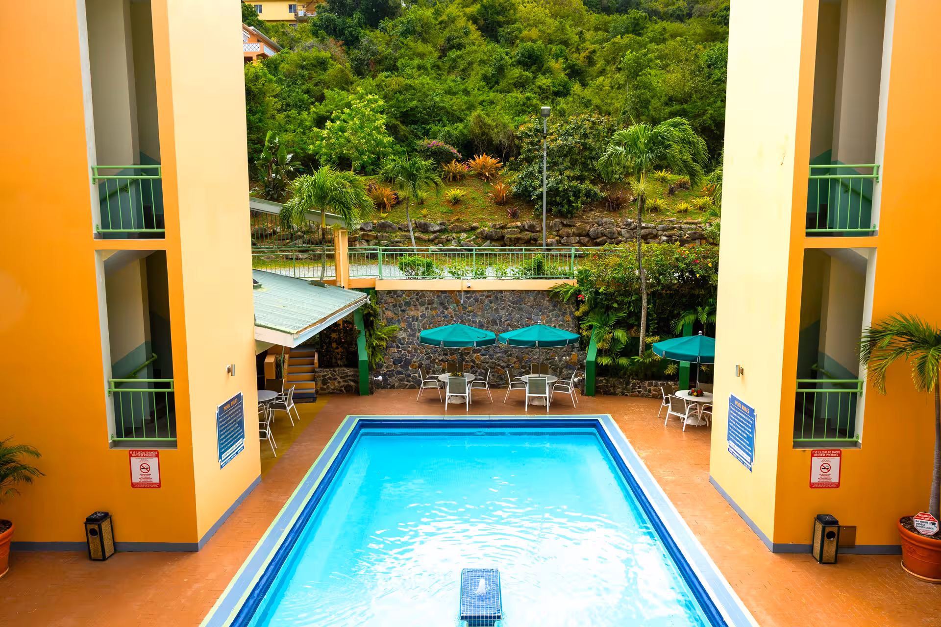 Outdoor rectangular swimming pool with turquoise water surrounded by tables with green umbrellas, chairs, and orange walls with green railings, set against a lush green hillside.