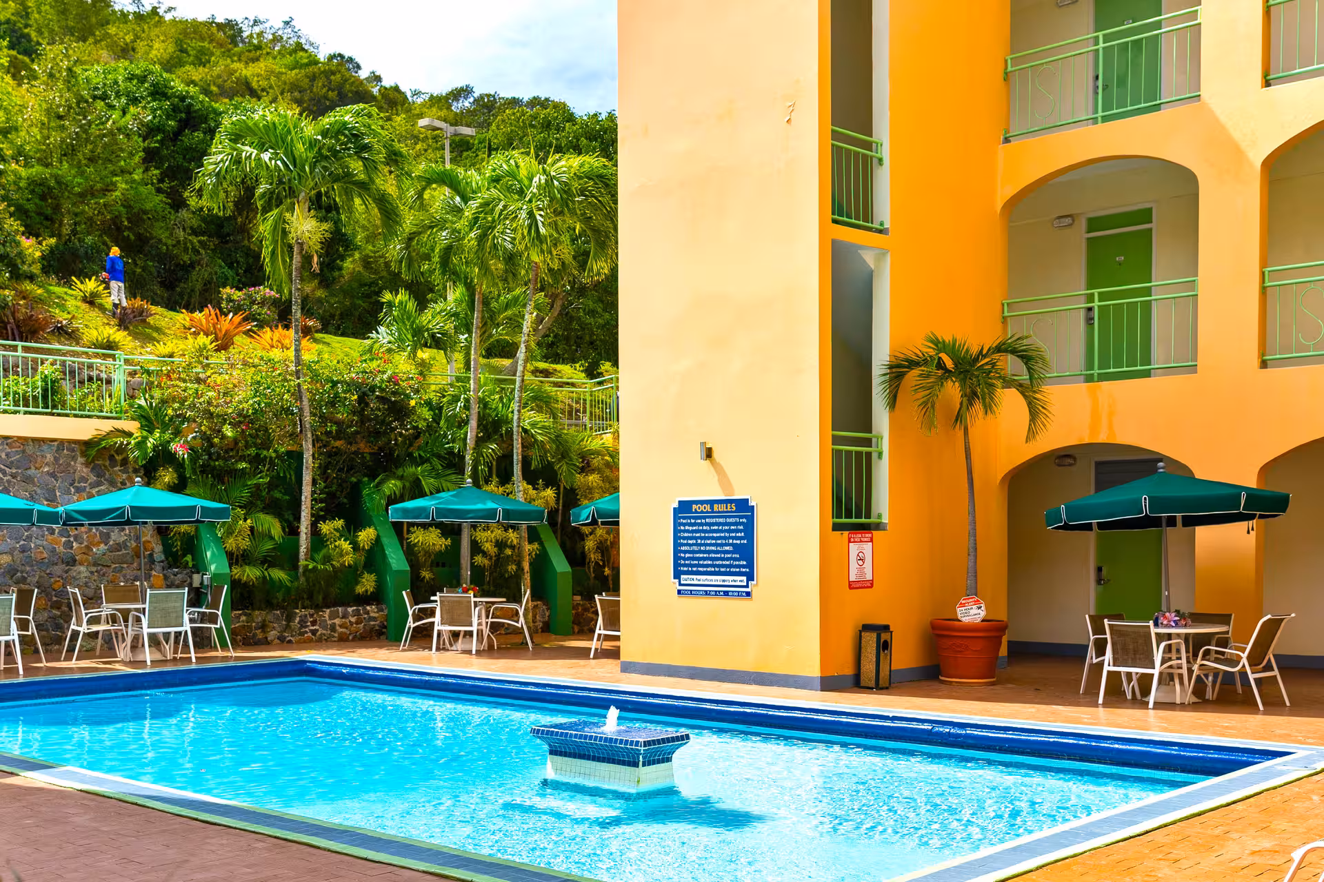 Outdoor swimming pool with blue water, surrounded by tables with green umbrellas and a yellow building with green balcony railings.