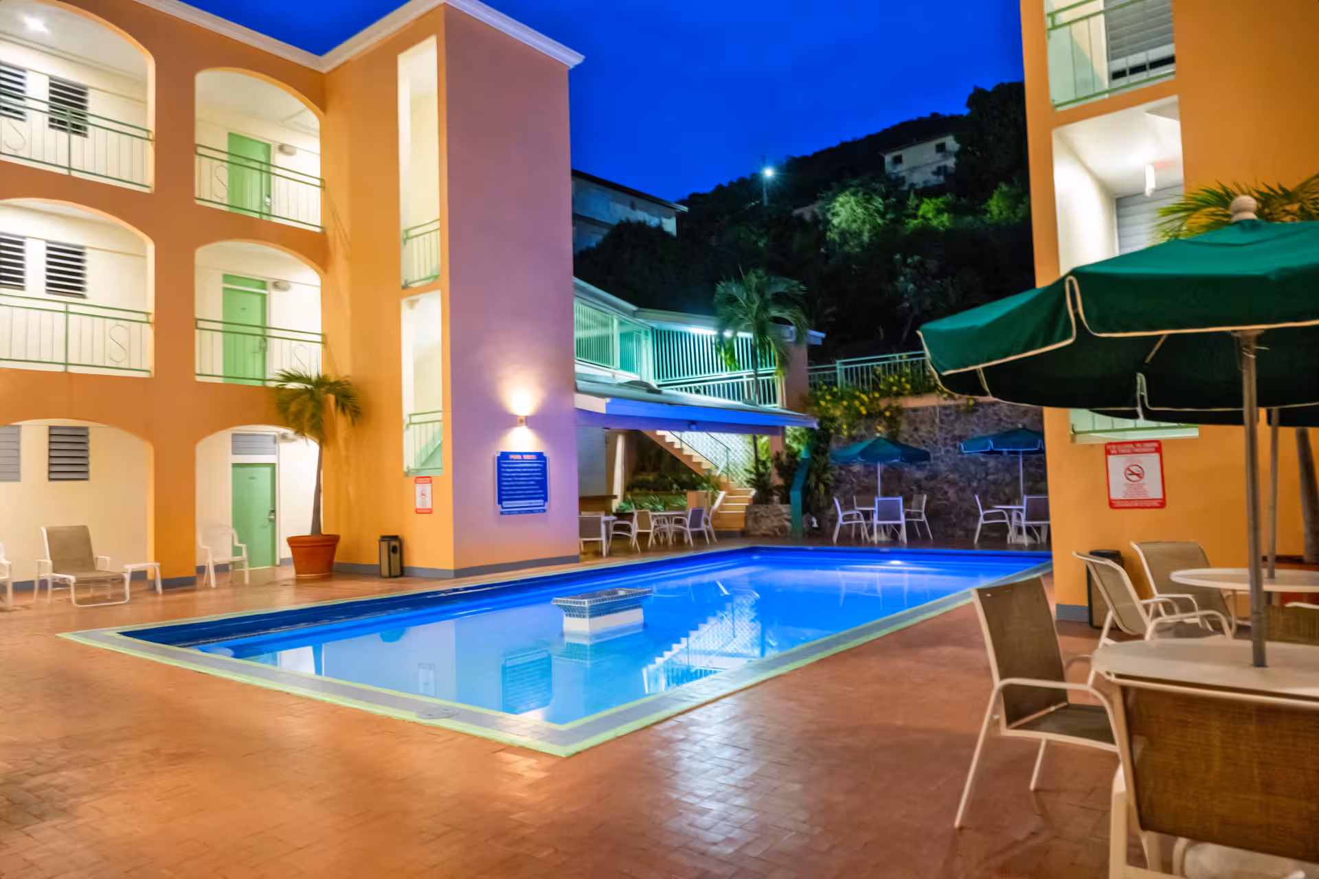 Outdoor swimming pool at a hotel courtyard at night with surrounding lounge chairs and green umbrellas.