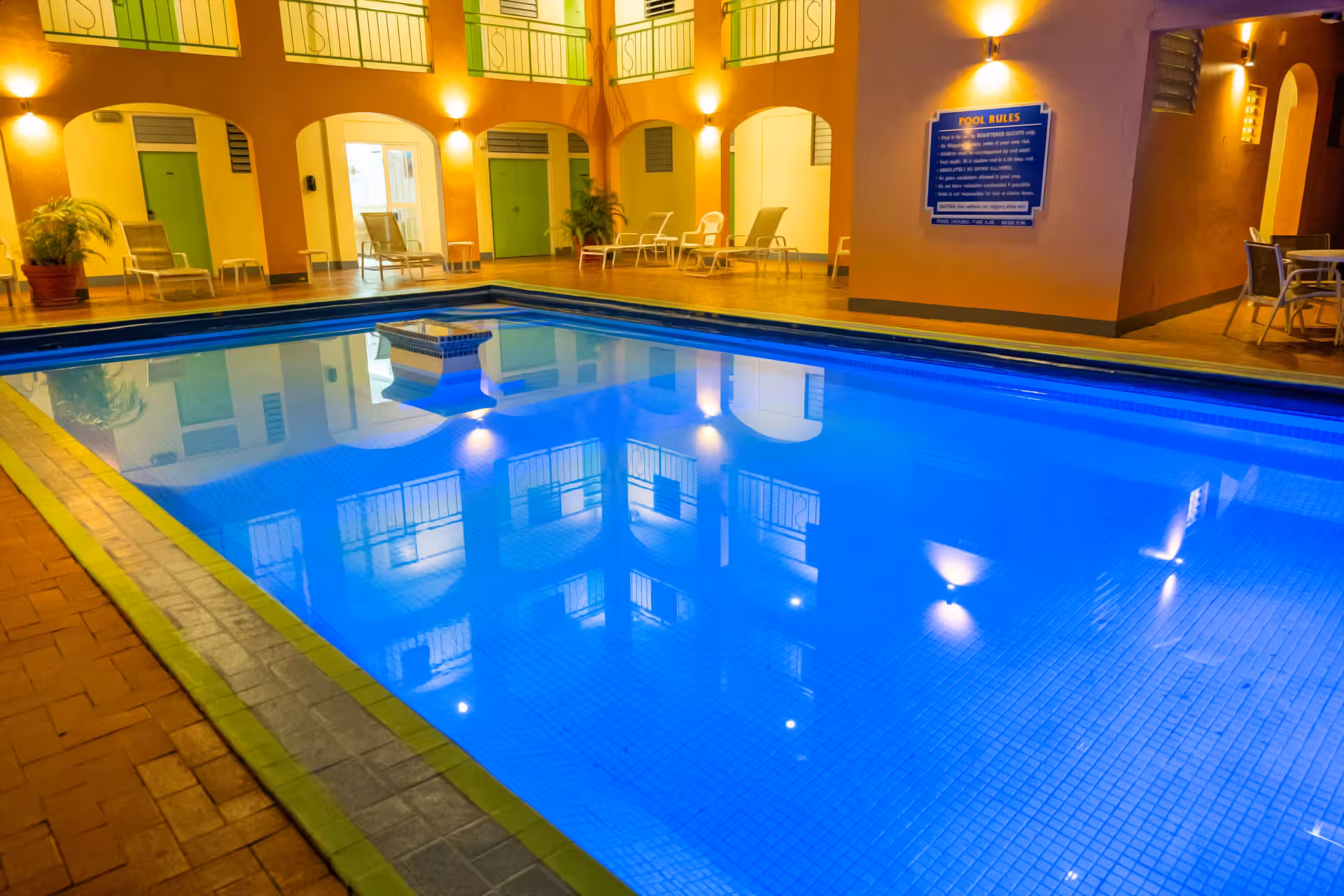 Indoor swimming pool illuminated with blue lighting surrounded by lounge chairs and warm yellow walls with green doors.